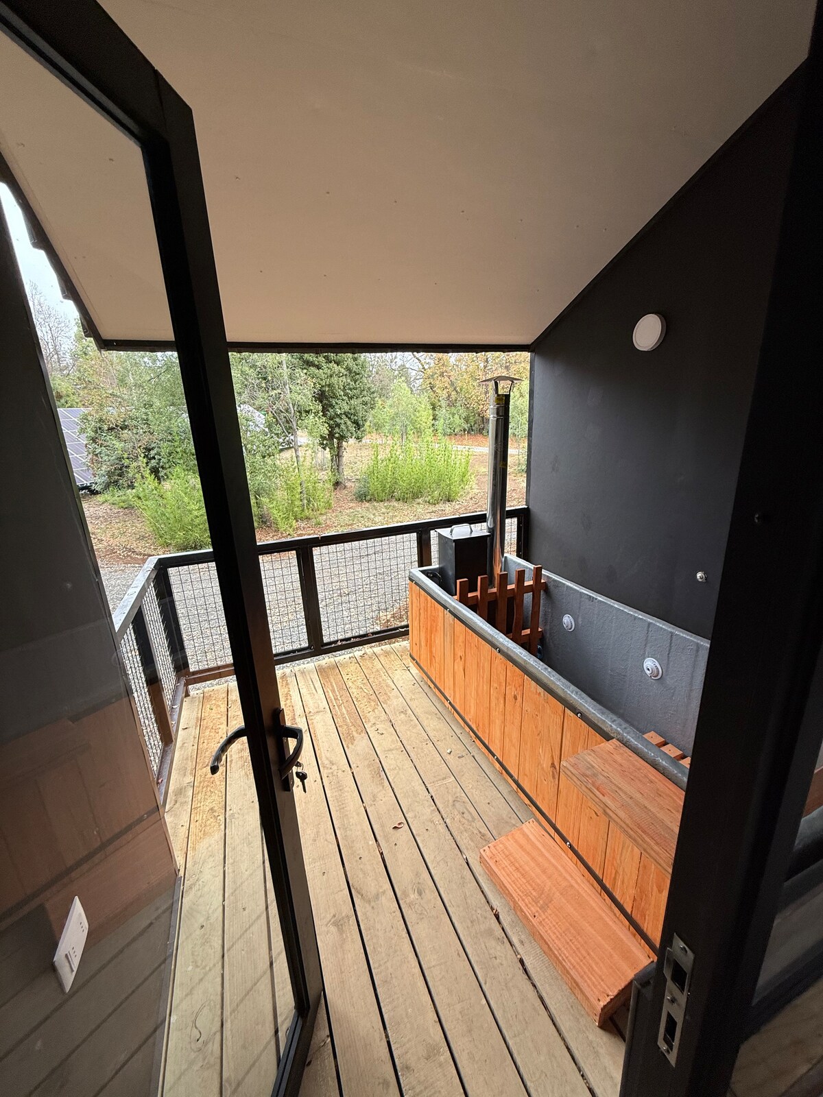 A private terrace features a small, wooden hot tub surrounded by natural greenery. The entrance is marked by glass doors that open onto a wooden deck, with a simple railing providing safety and open views of the tranquil surroundings.