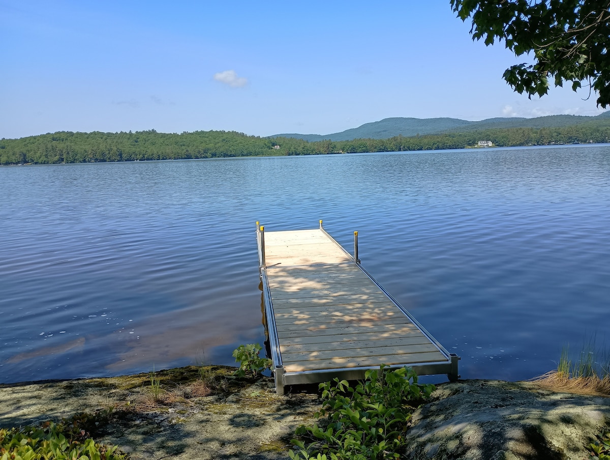 An aluminum dock extends gracefully over the calm surface of Lake Anasagunticook, surrounded by lush greenery and gently rolling hills in the background. The clear blue sky reflects on the water, creating a serene setting for relaxation and outdoor activities.