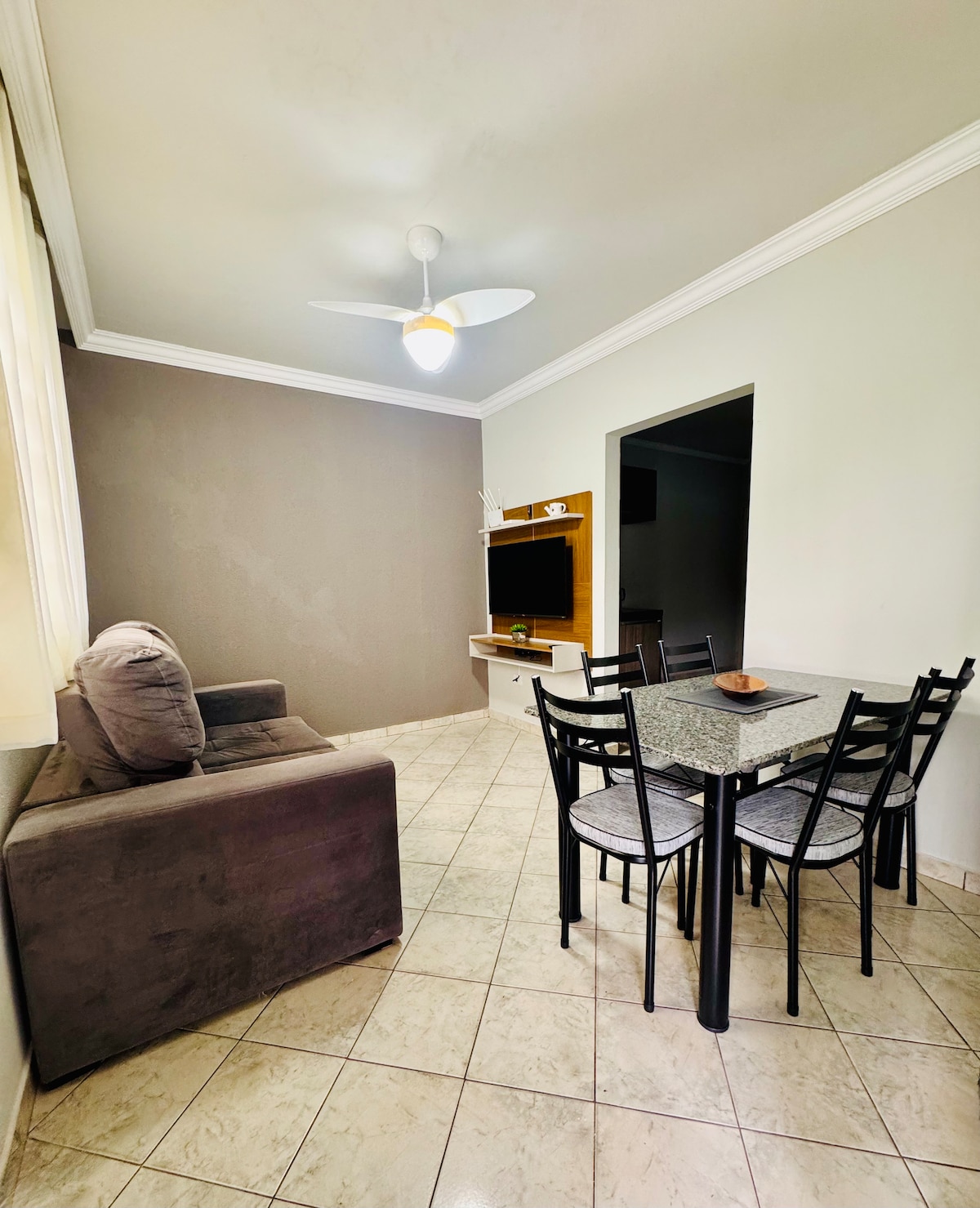A compact living area features a dark sofa and a dining table with four chairs. A ceiling fan provides airflow. The walls are painted in neutral tones, and a TV stand is visible against the wall, enhancing the functional layout of the space.
