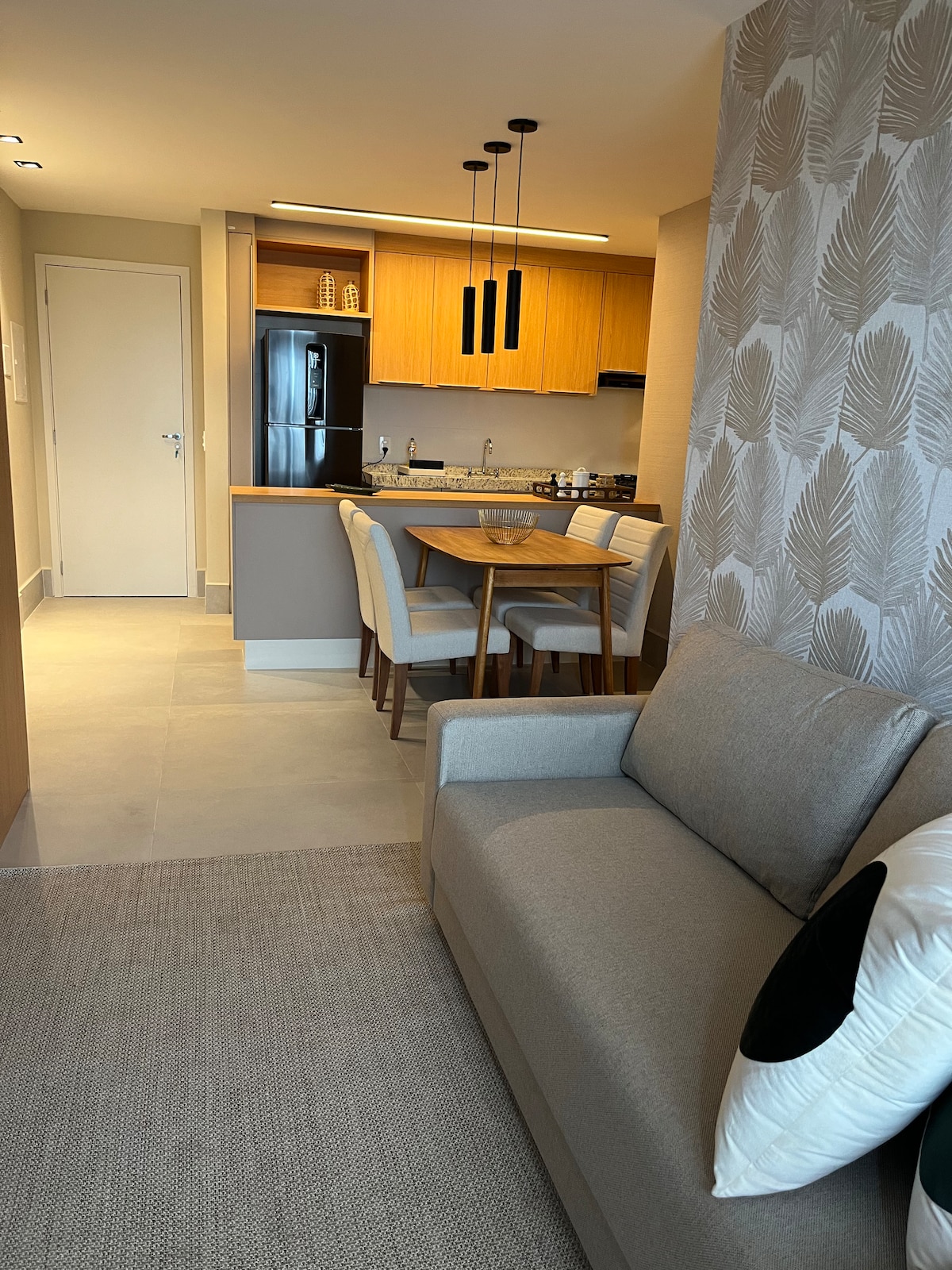 A modern kitchen area features wooden cabinetry and stainless steel appliances. A dining table with four chairs complements the open layout. A gray sofa with a decorative pillow is situated in the foreground, providing a welcoming space that connects with the kitchen and dining area.