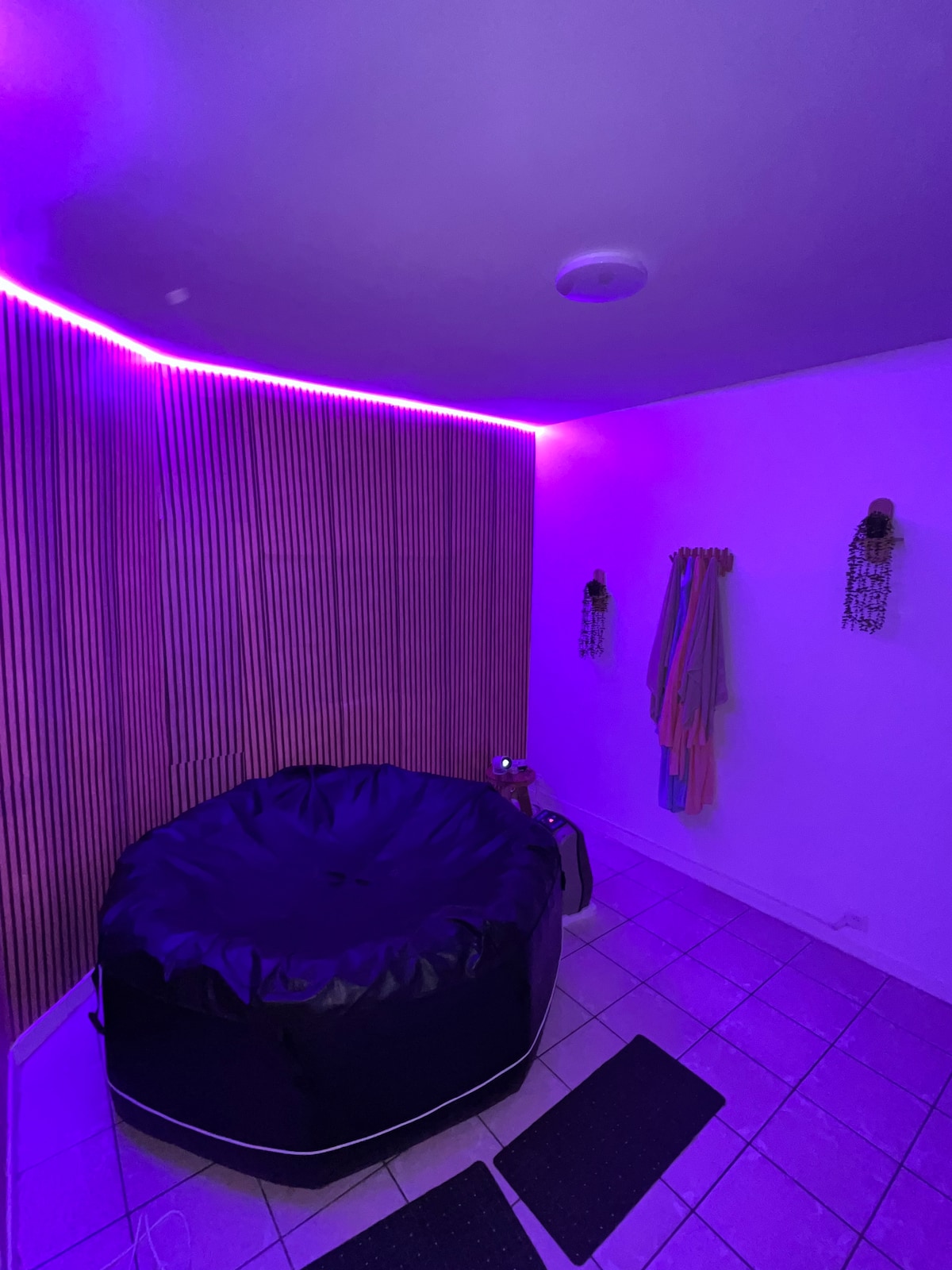 A cozy wellness space features a four-person jacuzzi illuminated with soft purple lighting. Textured wood panels are visible on one wall, while hanging towels add a touch of color. The tiled floor provides a clean and modern feel to the inviting area.