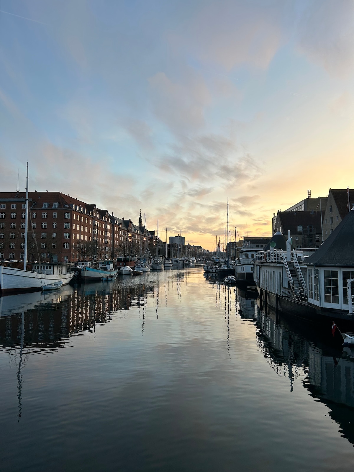 A tranquil canal scene is depicted at sunset, with gentle reflections of historic buildings and boats on calm water. The sky displays a gradient of soft colors, creating a serene backdrop for the lined vessels and architectural features along the waterfront.