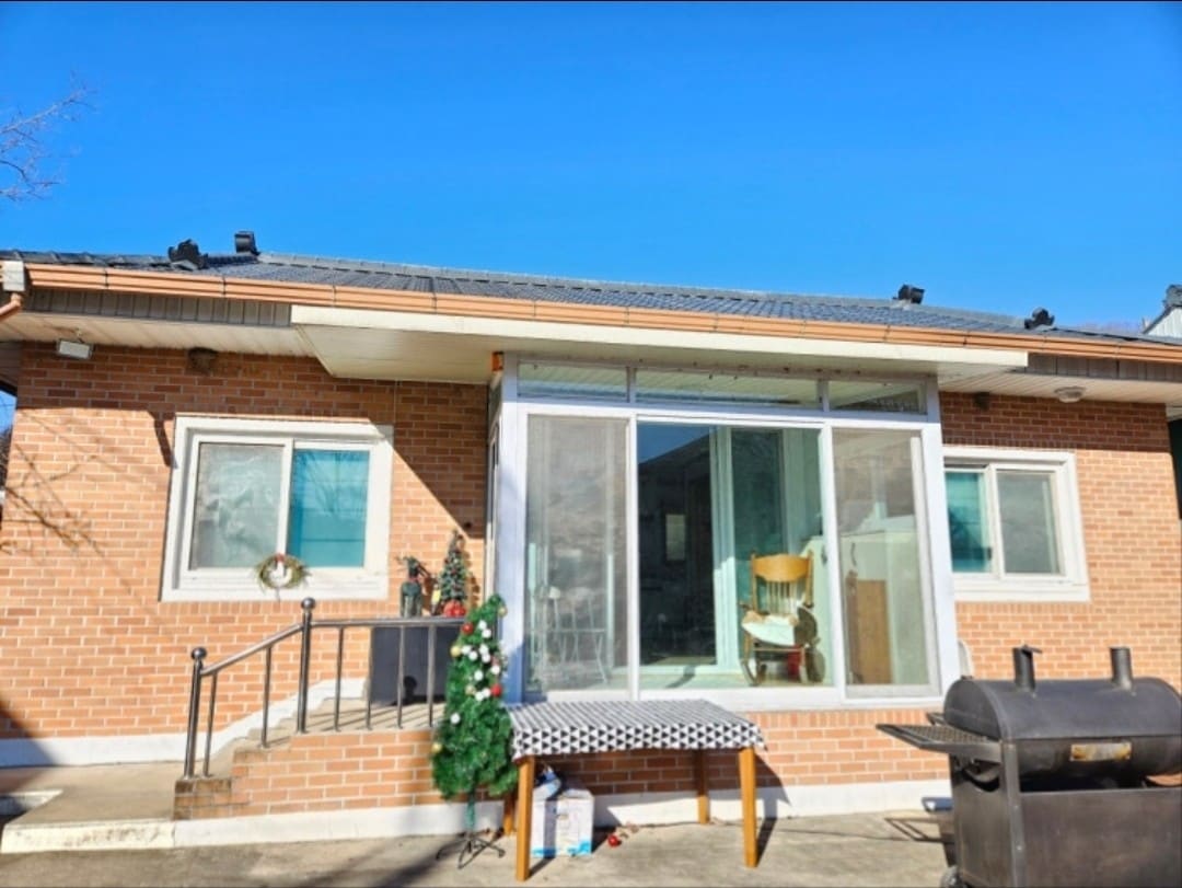 A brick exterior of a single-story house is showcased against a clear blue sky. Large windows allow natural light to enter, while a decorative tree stands next to the entrance. A barbecue grill is positioned on the patio, enhancing outdoor dining options.
