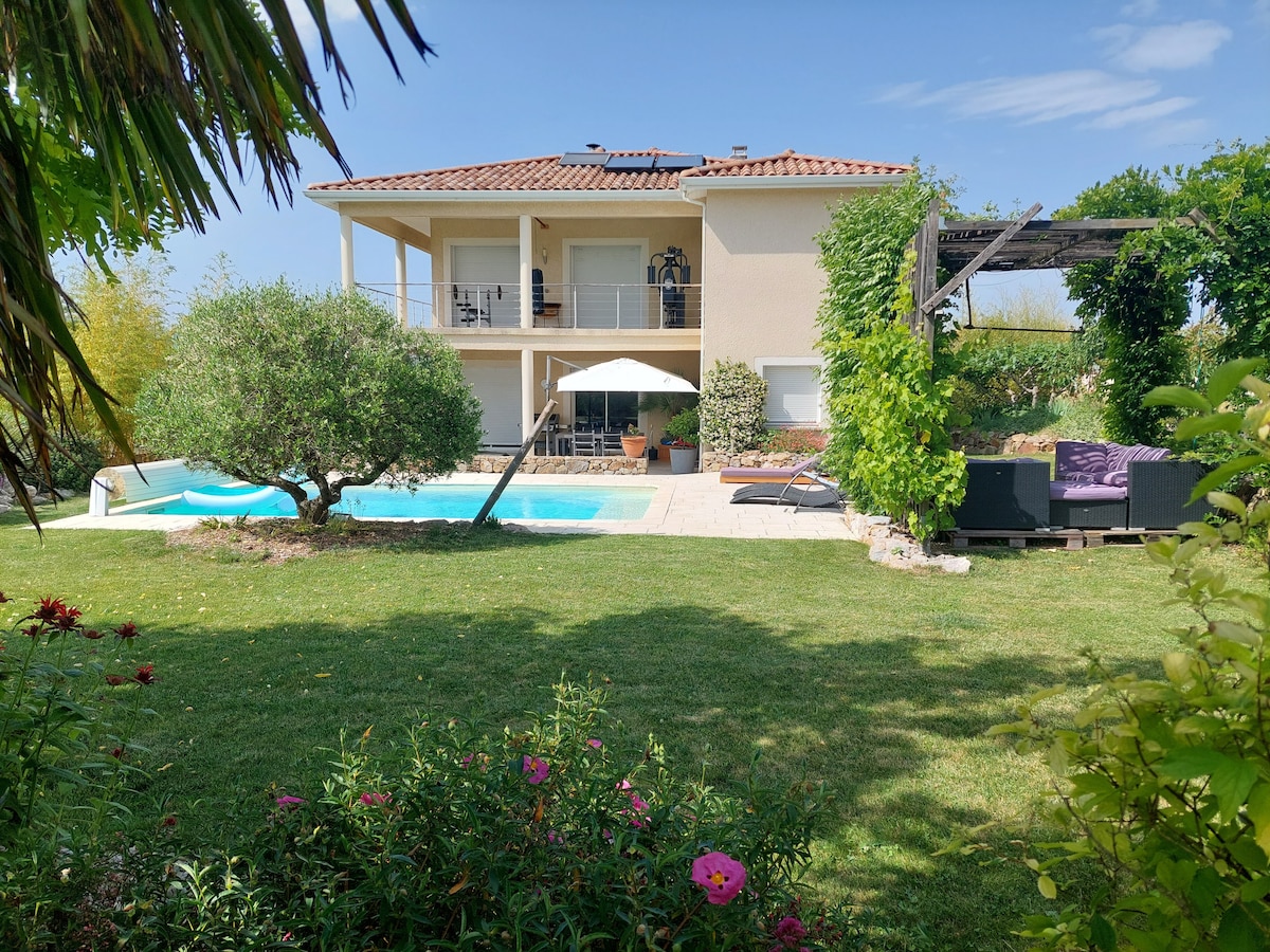 A spacious residence is set amidst a lush garden, featuring a landscaped lawn and a tranquil pool. The upper balcony is shaded with an umbrella, while comfortable seating is arranged nearby, inviting relaxation. Sunlight highlights the well-maintained greenery, creating a serene outdoor atmosphere.