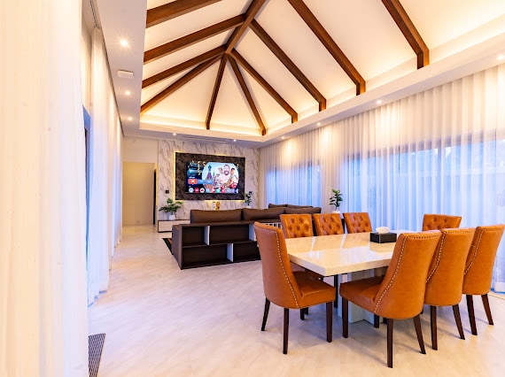 A spacious dining area features a large marble table surrounded by eight upholstered chairs. A stylish sofa set is positioned in front of a wall-mounted television. Soft natural light filters through sheer curtains, illuminating the room's modern design and distinct ceiling beams.