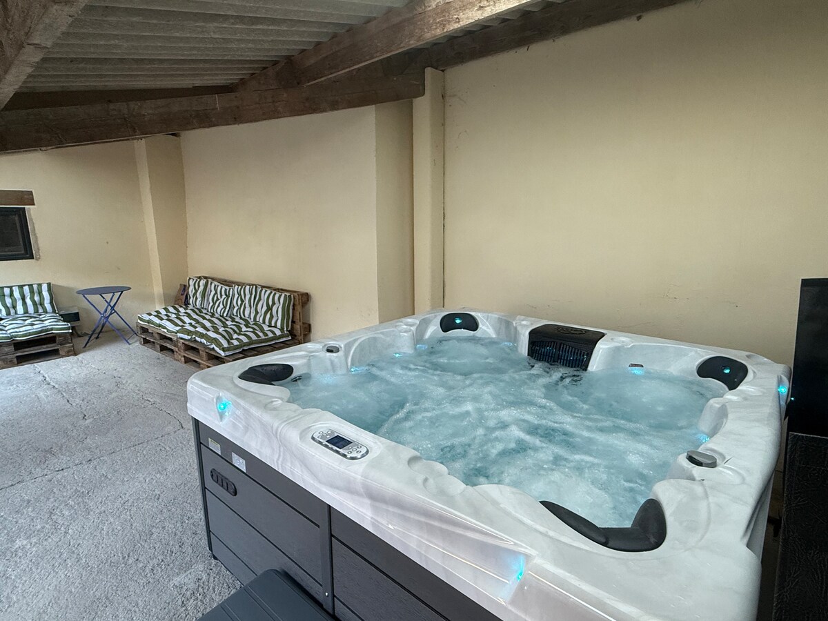 A spacious hot tub is located indoors, featuring swirling water and built-in jets. Nearby, a casual seating area with striped cushions is available, offering a relaxed space to unwind. The ceiling beams add character to the room, contributing to a cozy atmosphere.