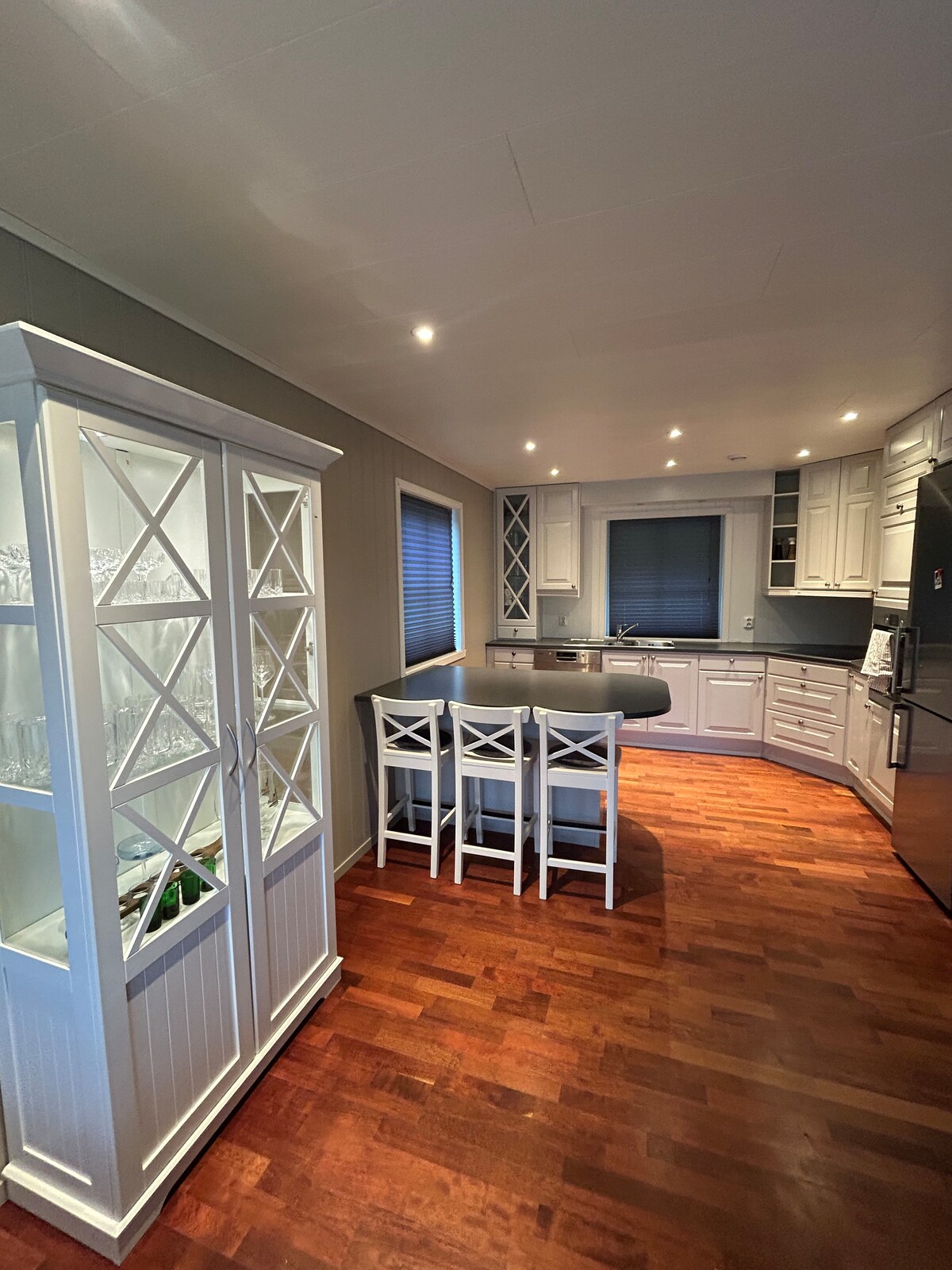 The kitchen features a modern design with white cabinetry and sleek dark countertops. A glass-front cabinet displays glassware, while a central island offers seating for four. Ample lighting illuminates the space, complemented by warm wood flooring and a window with blinds.