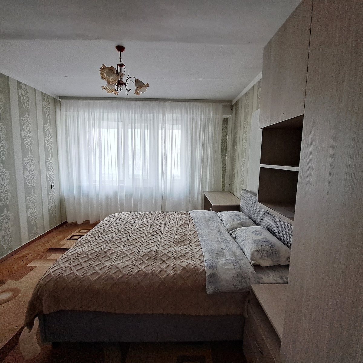 A sleeping area features a spacious bed with a patterned blanket, complemented by light-colored bedding. A large window is dressed with sheer curtains, allowing natural light to fill the room. A wooden dresser is positioned next to the bed, enhancing the room's functionality.