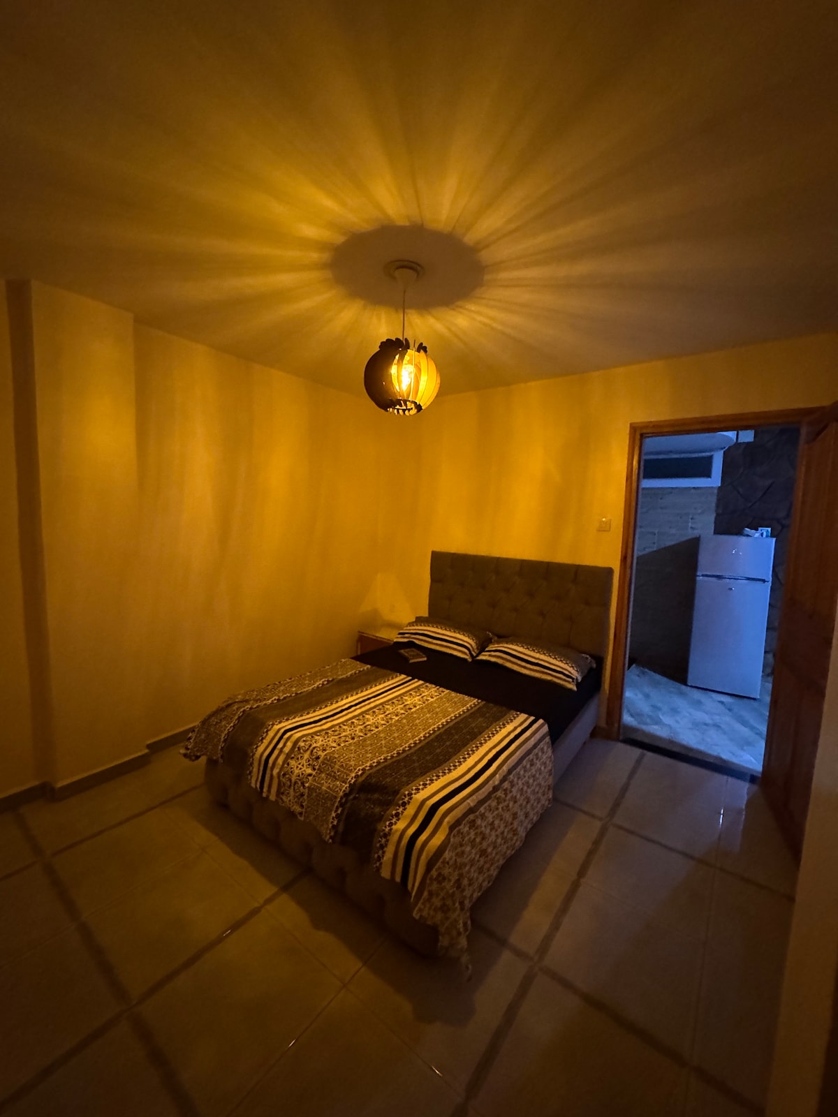 A cozy sleeping area is depicted with a spacious bed adorned with a patterned blanket. Soft lighting is provided by a lamp, casting warm tones on the walls. The room features tiled flooring and a glimpse of a kitchenette in the background.