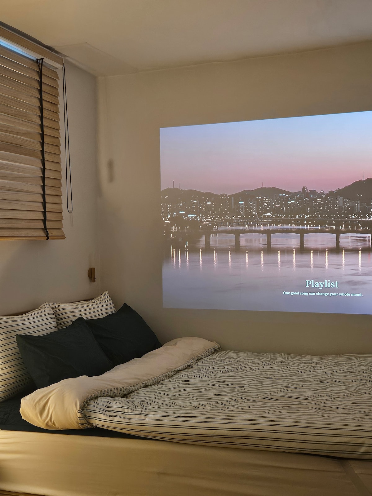 A cozy sleeping area features a spacious bed with soft bedding in neutral tones. A soft light projects a scenic view of the city skyline onto the opposite wall, creating an inviting atmosphere. Natural light filters through the wooden blinds, enhancing the relaxed setting.