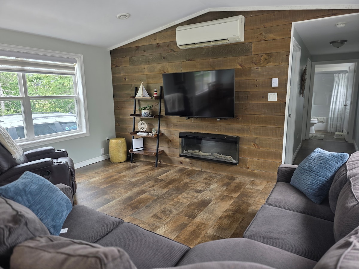 A cozy living area features a wooden accent wall and a flat-screen television mounted above an electric fireplace. Two plush sofas, one in a dark tone and one with blue accent pillows, are arranged for comfort, while natural light floods in through a nearby window.