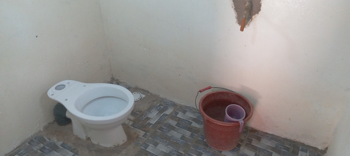 A simple bathroom is depicted, featuring a modern toilet alongside a bucket filled with water and a small cup. The tiled floor provides a clean and functional space, while the light wall color enhances visibility.