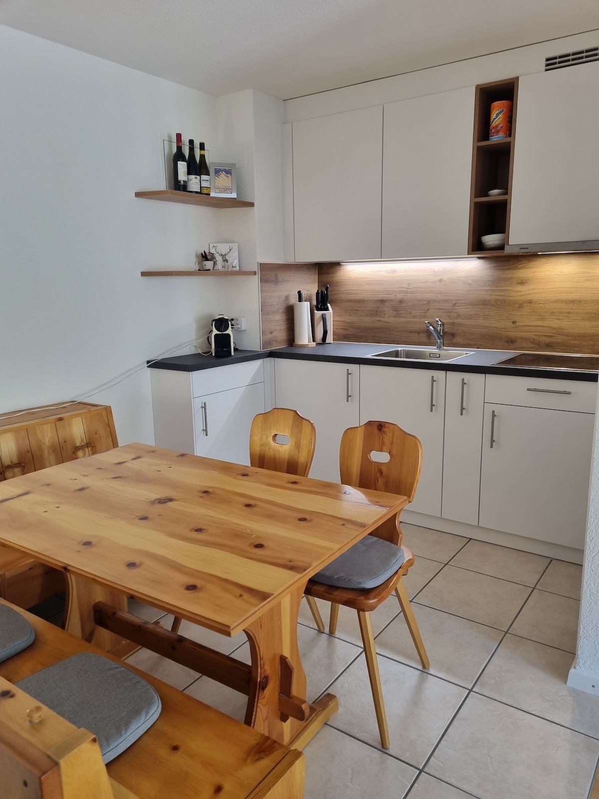 A compact kitchen area features light wooden shelves displaying wine bottles and decorative items. A sleek countertop with modern appliances is complemented by a wooden dining table and two chairs. Bright, ambient lighting highlights the stylish cabinetry and creates a warm space.