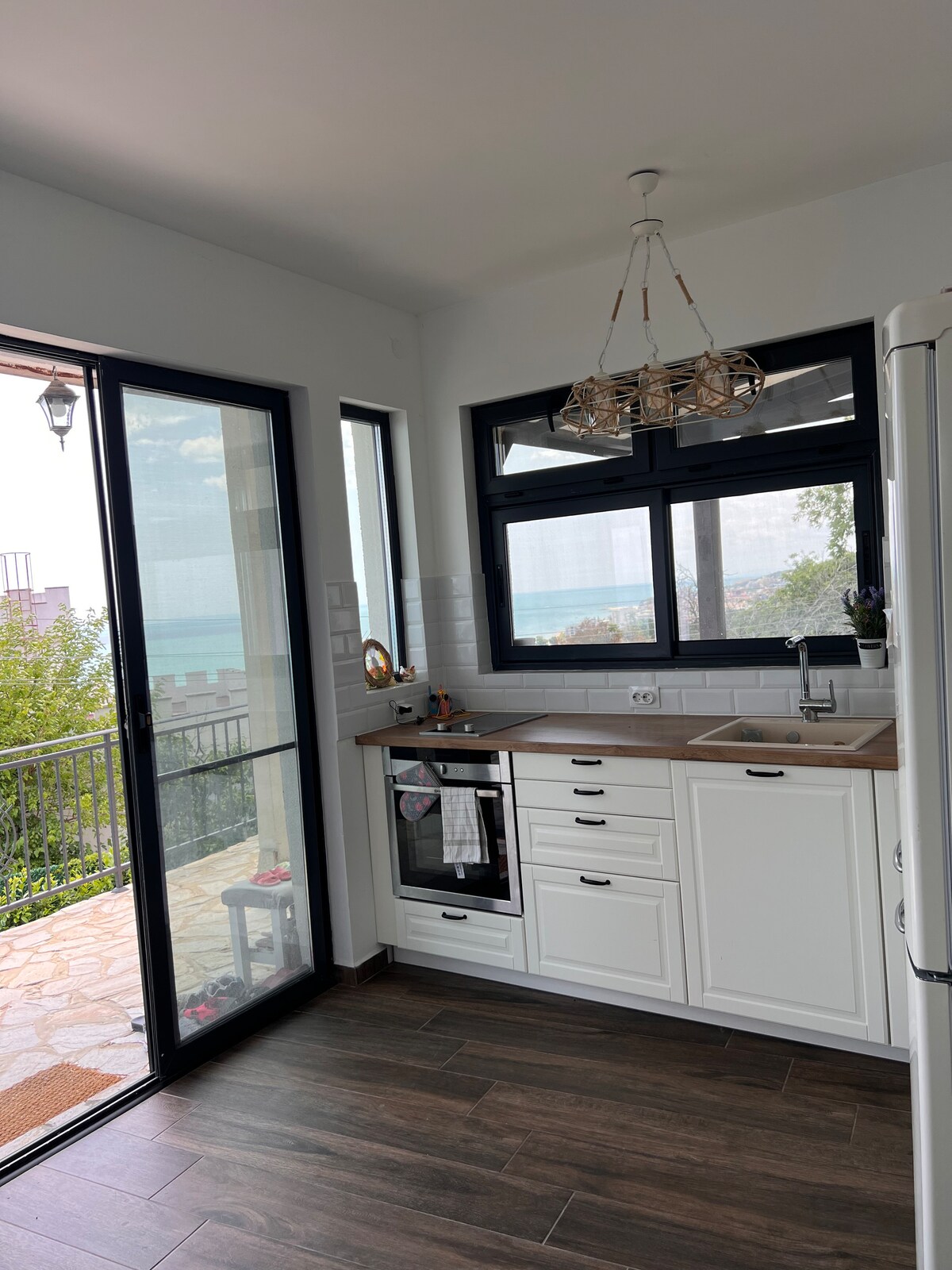 A modern kitchen features white cabinetry and a wooden countertop. Large windows allow natural light to fill the space, offering views of the sea. A sleek stove and a vintage-style refrigerator are visible, enhancing the functional yet welcoming atmosphere.