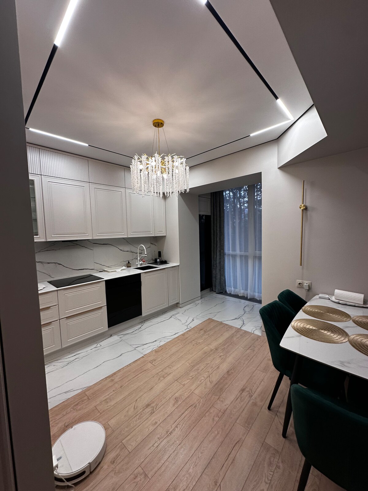 A contemporary kitchen features light cabinetry paired with sleek countertops and modern appliances. A stylish chandelier hangs from the ceiling, illuminating the space. An adjacent dining area displays a set of plates on a table surrounded by dark green chairs, enhancing the overall dining experience.