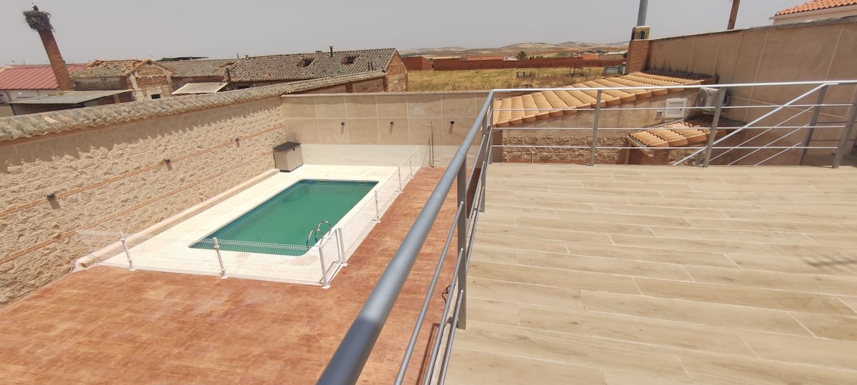 An outdoor terrace provides a view of the rectangular pool surrounded by a tiled deck. A spacious area features a wooden awning, with the backdrop of stone walls and farmland in the distance. The serene atmosphere is enhanced by clear blue skies.