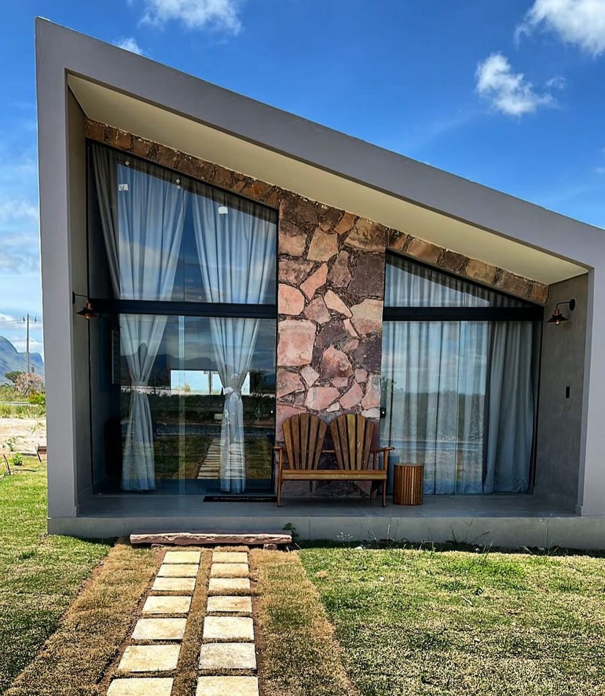 The exterior of a modern cabin is showcased, featuring a distinctive sloped roof and a natural stone accent wall. Large glass windows are framed by light, flowing curtains. A wooden chair and side table contribute to a relaxed outdoor atmosphere, with a pathway lined by stone pavers leading to the entrance.
