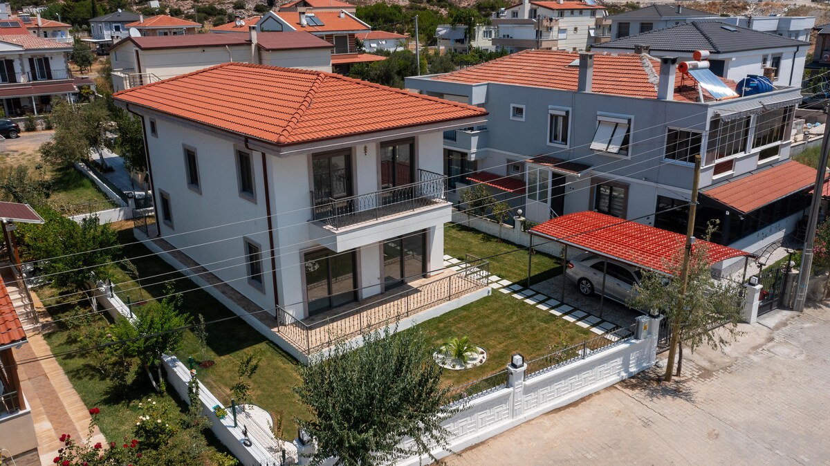 The two-story house features a neatly landscaped front yard, surrounded by a white fence. A spacious patio area is visible, providing access to the main entrance. Nearby, another modern structure and parking spaces complement the residential setting.