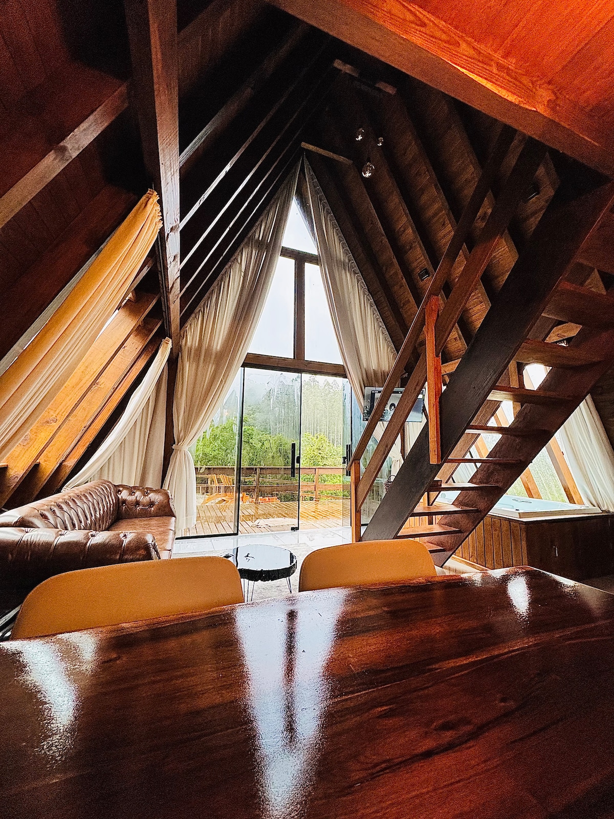 A spacious interior showcases tall, angled wooden beams against large windows that invite natural light. A plush seating area is complemented by a dark coffee table, while the wooden dining table offers a perfect setting for intimate meals. Sheer curtains drape gracefully, adding softness.