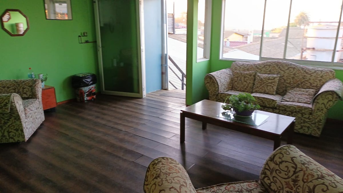 A spacious living area is presented with vibrant green walls and large windows, allowing natural light to fill the space. Two patterned armchairs and a matching sofa are arranged around a wooden coffee table, which is adorned with a potted plant. A door leads to a balcony.