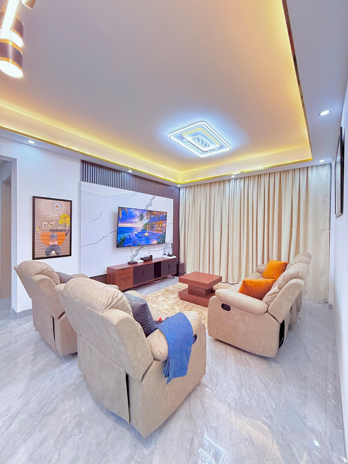 A spacious living area features two reclining sofas adorned with orange cushions. A large television is mounted against a feature wall, while soft, ambient lighting enhances the neutral décor. Floor-to-ceiling curtains provide a sense of privacy and elegance.