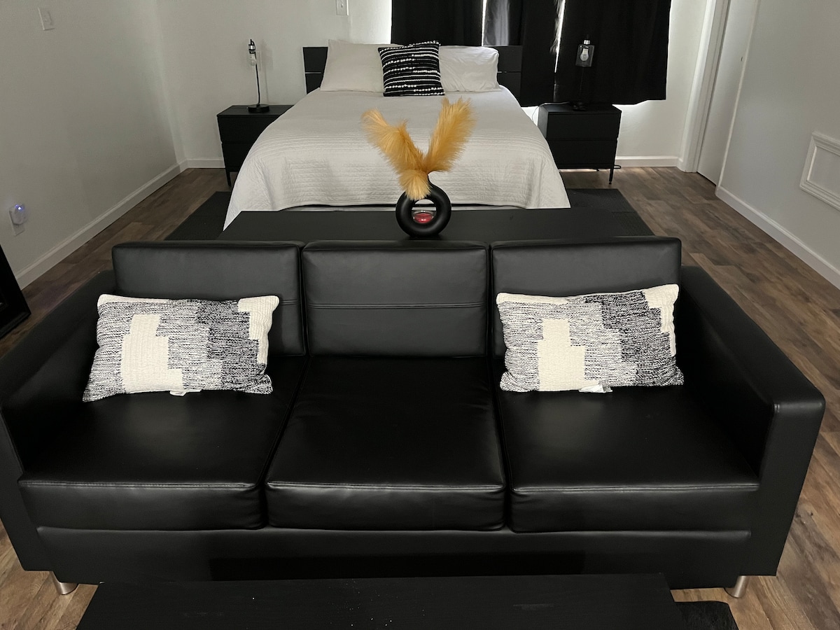 A modern sofa with dark leather upholstery is positioned in the foreground, complemented by decorative pillows. The spacious bed with neutral bedding is visible in the background, along with matching bedside tables. Natural light filters through dark curtains, creating a serene atmosphere.