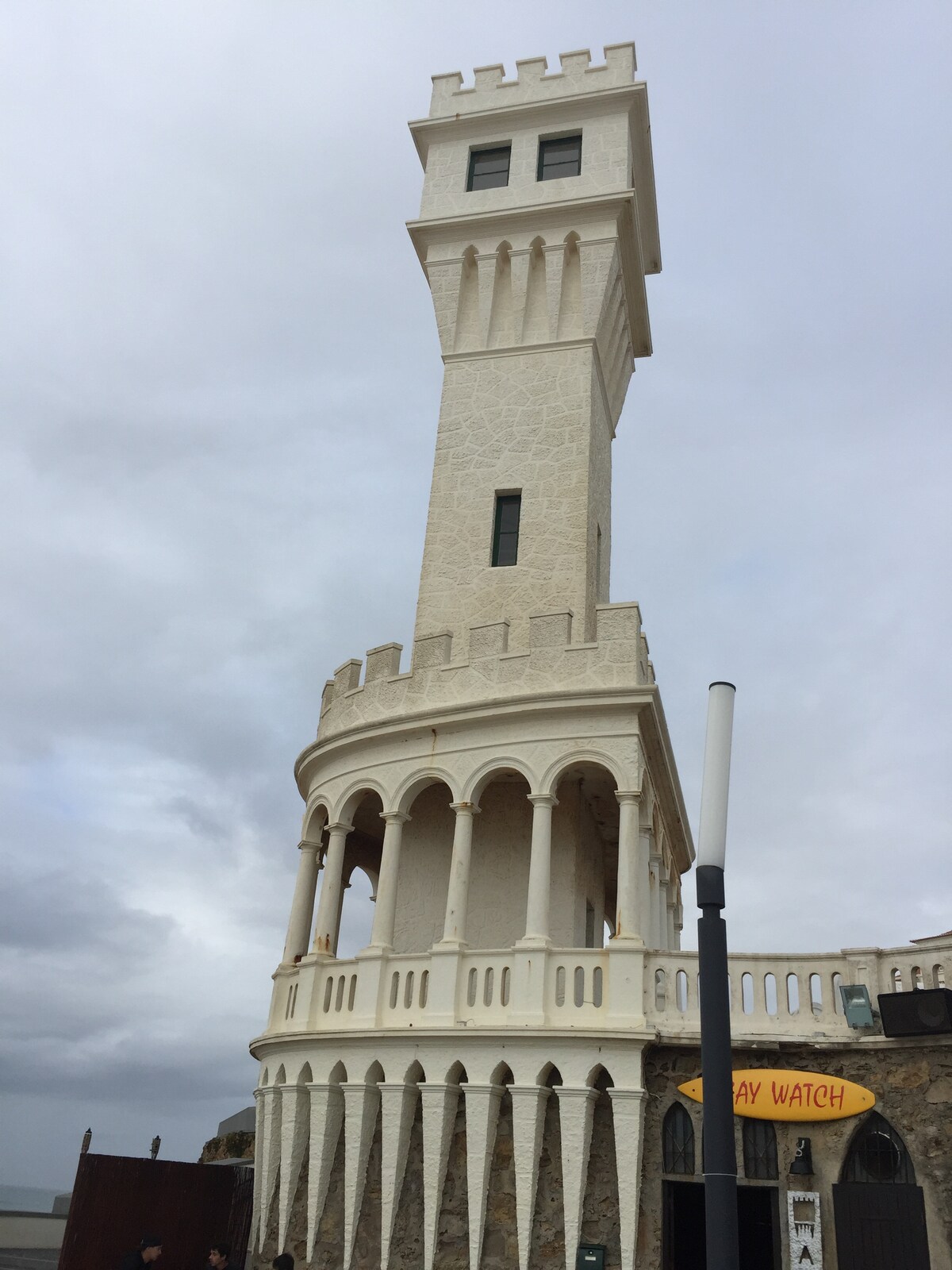 An architectural structure features a tall, turret-like tower with a crenelated roof and arched windows. The building's stone façade displays intricate details, while a sign reading 'Lifeguard' is visible at the base. Overcast skies provide a muted backdrop.