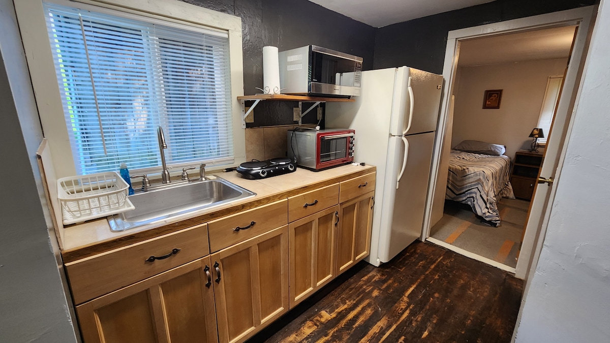 A compact kitchen area is presented, featuring wooden cabinetry and a stainless steel sink under a window. Appliances include a microwave and a refrigerator. A door leads to a cozy sleeping area with a neatly made bed visible in the background.