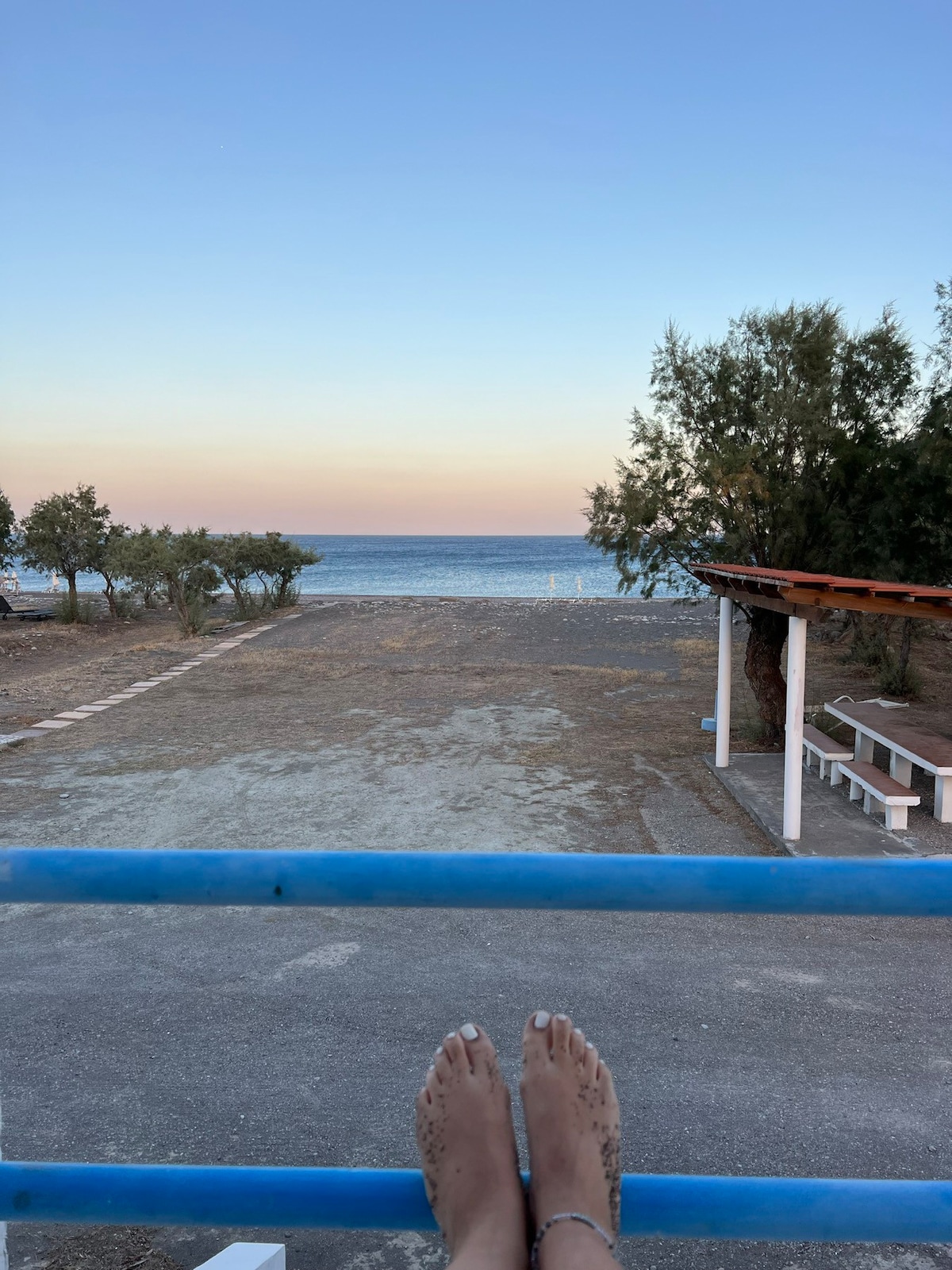 A serene coastal view is presented, featuring a calm sea and clear skies. The horizon transitions into soft pastel colors. In the foreground, a shaded seating area with a wooden roof is visible, along with scattered trees framing the landscape.