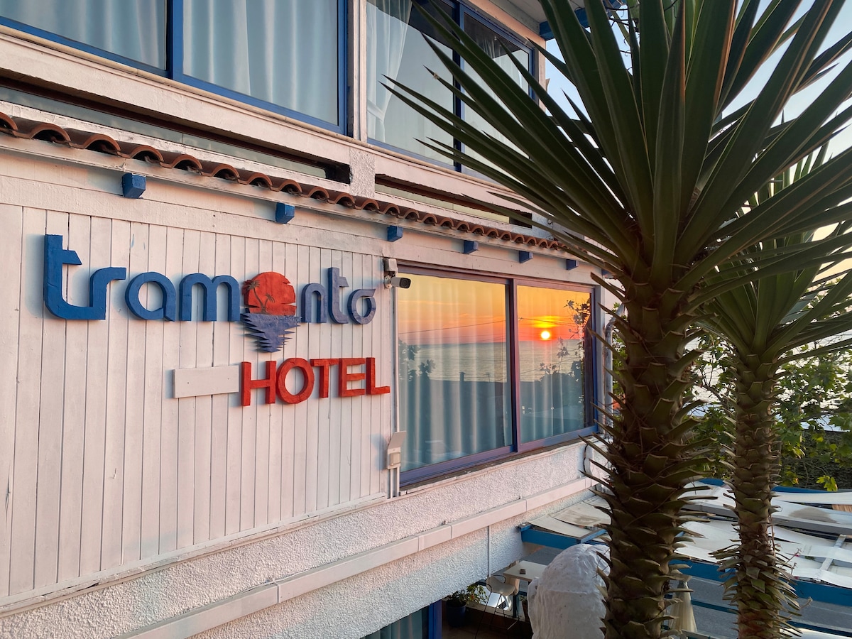 The exterior of the hotel is depicted with a palm tree nearby. The hotel name is visible on the building, with sunset reflections in the glass windows, creating a serene coastal atmosphere.
