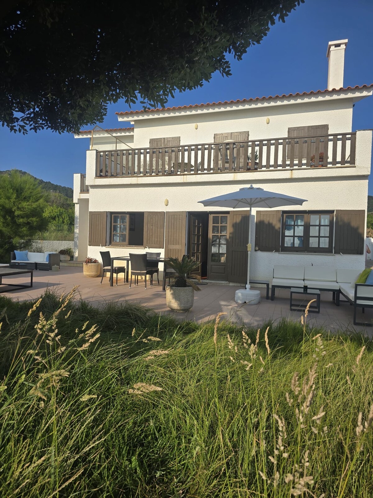 A two-story house is surrounded by lush green grass and features a balcony with wooden railings. The patio area is furnished with outdoor seating and a shade umbrella, providing a relaxing space. Nearby trees add to the tranquil setting in the background.