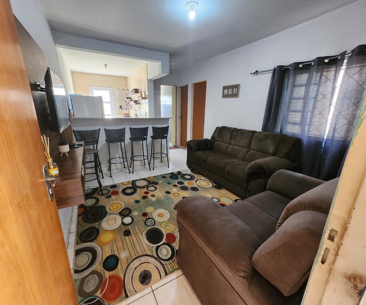 A living area features a dark upholstered sofa and recliner, arranged around a circular patterned rug. A television is mounted on a wooden stand, and a bar area with high stools can be seen in the background, offering a connection to the kitchen space.