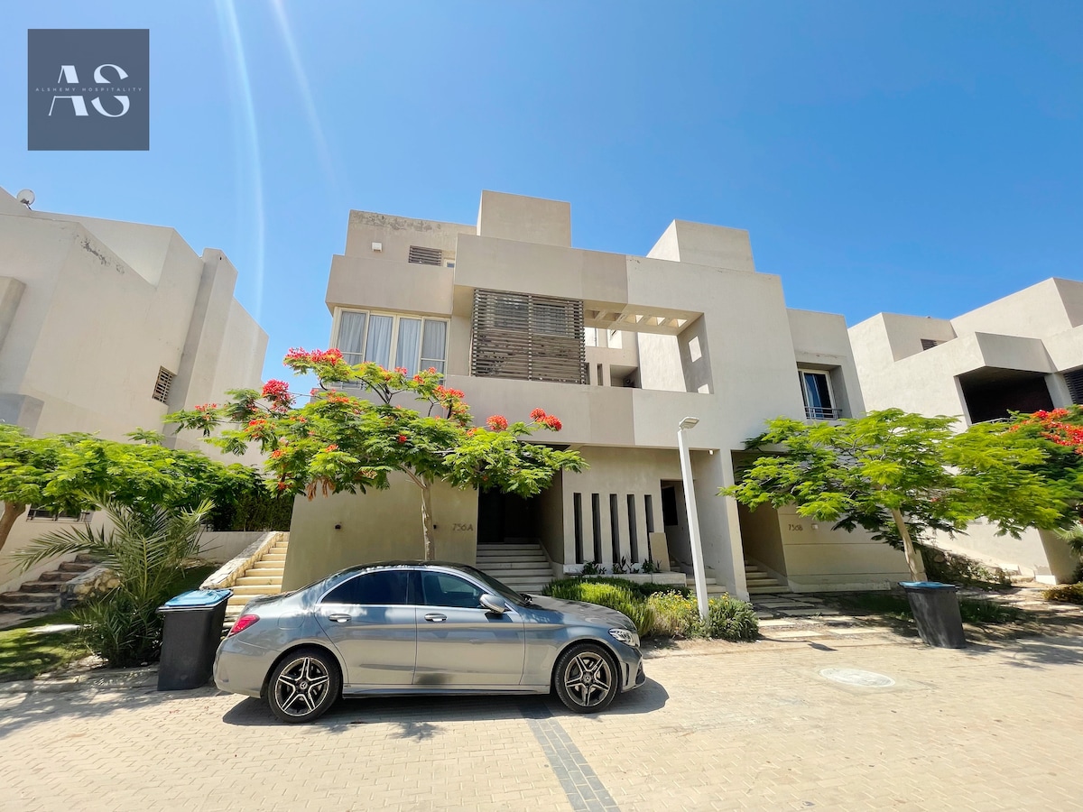 The image displays a modern architectural structure featuring clean lines and a neutral color palette. A silver car is parked in front of the building, which is surrounded by lush greenery and flowering trees. Bright sunlight casts clear shadows on the paved driveway.