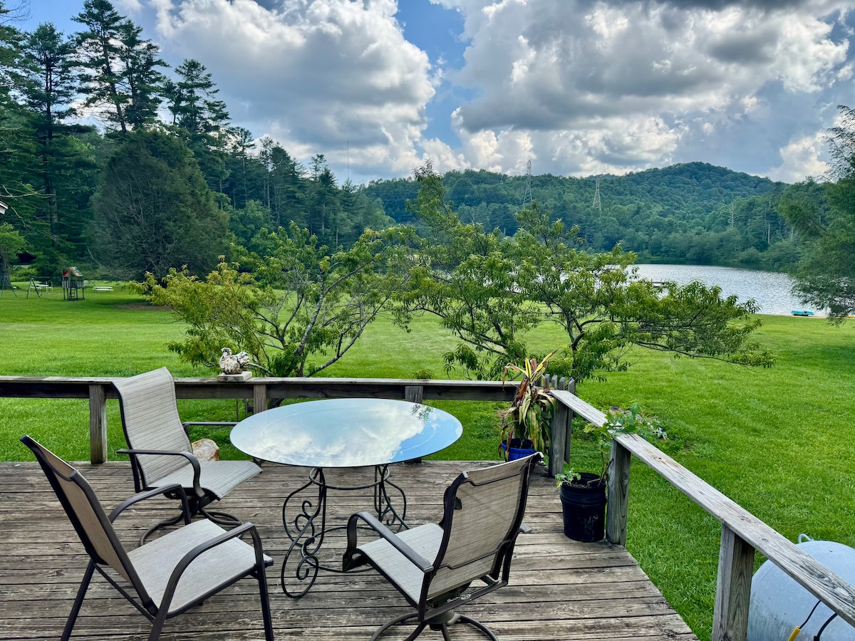 A wooden deck features a round glass table surrounded by two chairs. Lush, green grass extends towards a serene lake, framed by gentle hills and trees under a partly cloudy sky.