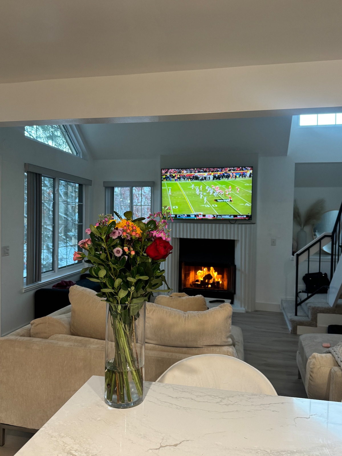 A dining table with a marble top is set in the foreground, featuring a tall vase filled with colorful flowers. In the background, a cozy living area is highlighted by a fireplace and a television displaying a sports game. Large windows allow natural light to fill the space.