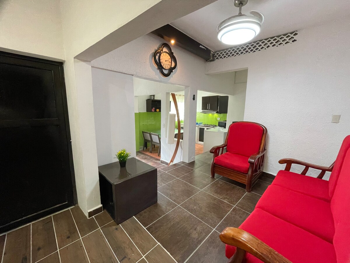 A welcoming living area is shown, featuring two comfortable red chairs and a small dark table. A clock hangs on the wall, and a green accent wall is visible leading into the kitchen area. Natural light filters in, enhancing the space's brightness.