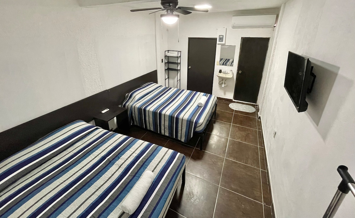 The guest room features two spacious beds, each covered with striped linens in blue and white tones. A ceiling fan is mounted above, and a wall-mounted TV is visible. A small sink and mirror area is seen in one corner, along with a black dresser.
