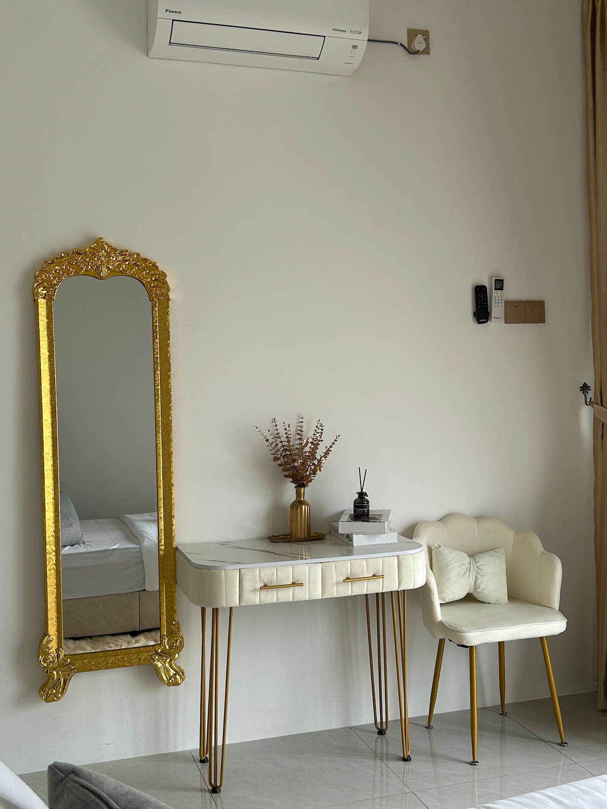 A vanity table with a white surface and gold legs is positioned beside a gold-framed mirror. A decorative vase with dried flowers and a small diffuser are placed on the table. A light-colored chair complements the setup, against a soft, neutral wall.