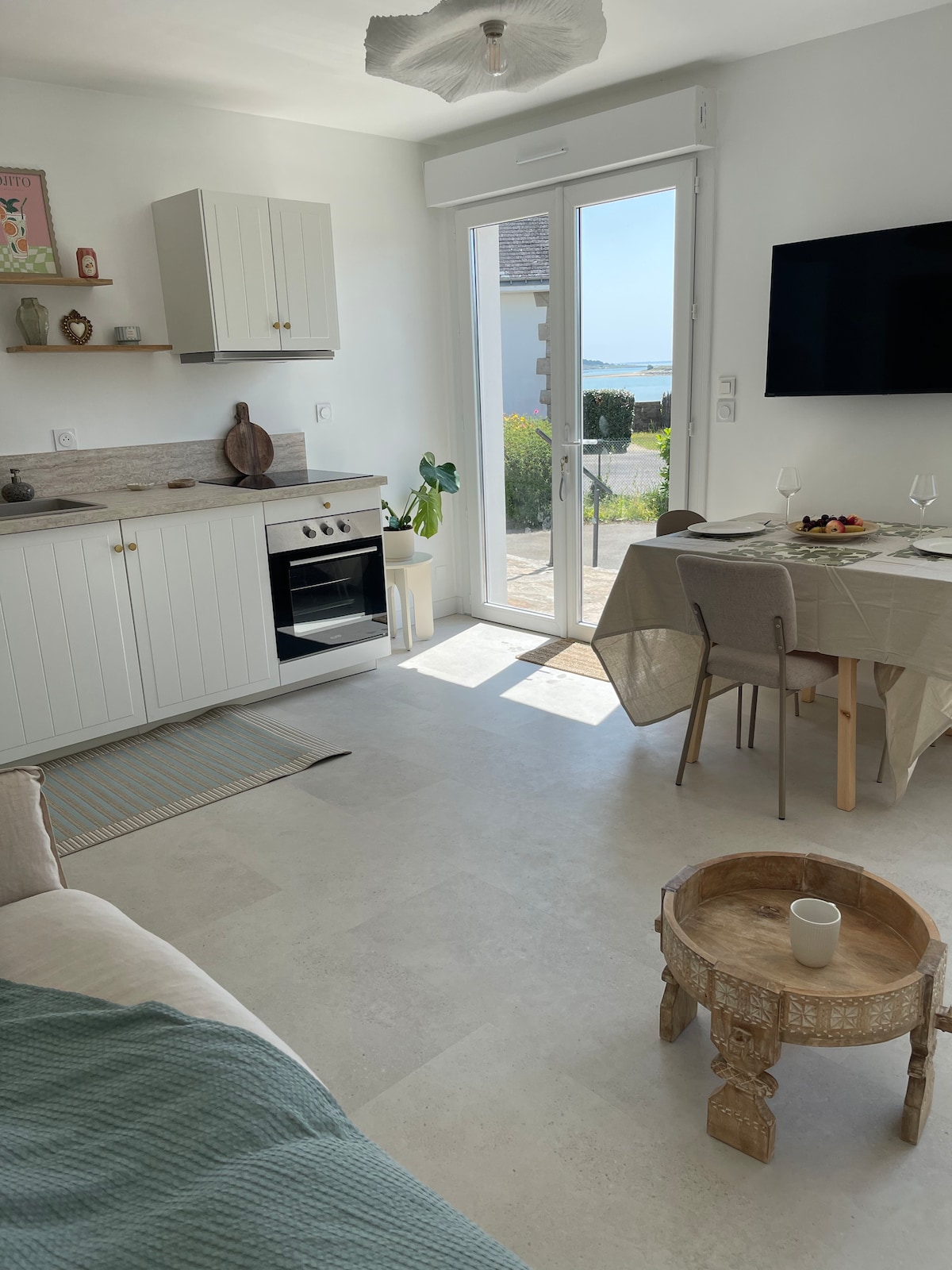 A bright living space showcases a kitchen area with white cabinetry and modern appliances, including an oven. A dining table for four and a cozy seating area are present, with sunlight streaming through large glass doors that open to a view of the sea and outdoor space.