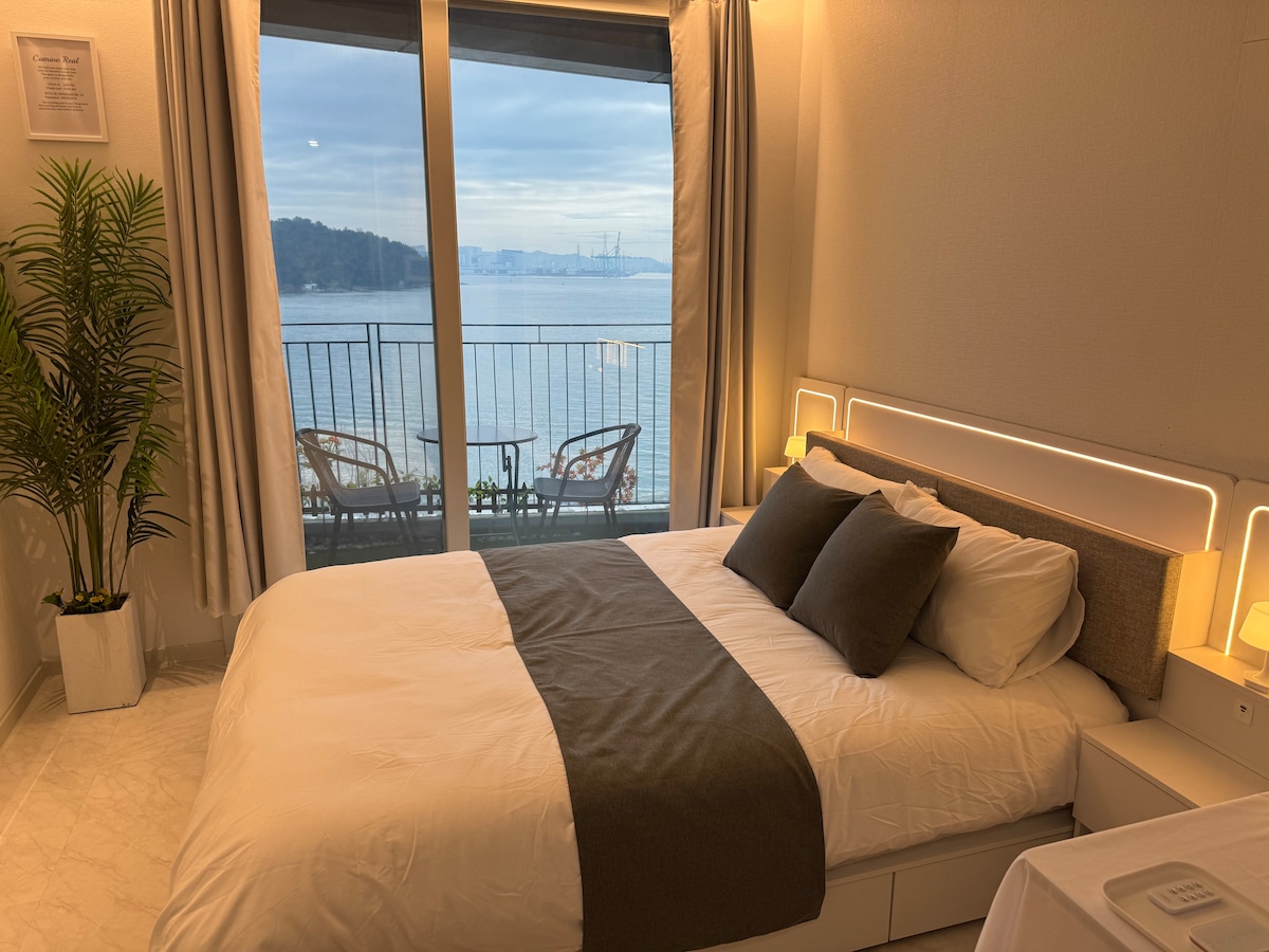 A spacious bed is dressed in neutral bedding, featuring complementary pillows. Large glass doors offer a sweeping view of the water and distant hills, while small outdoor seating is visible on the balcony. Soft lighting is provided by lamps on either side of the bed.