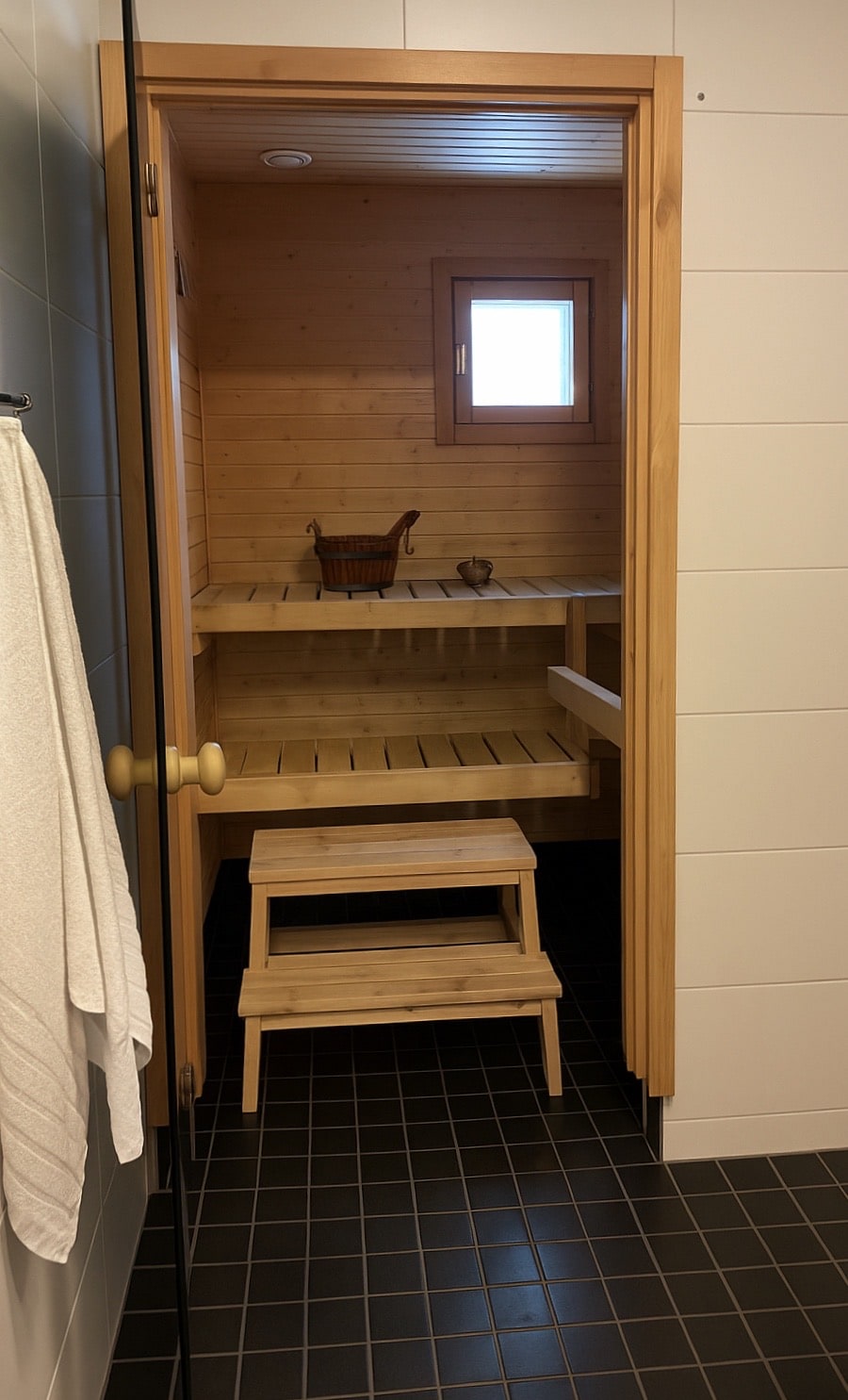 A wooden sauna is framed by light-colored wood paneling, creating a warm and inviting space. A small wooden stool is placed inside, and a decorative item rests on a shelf. A window allows natural light to enter, enhancing the serene atmosphere.