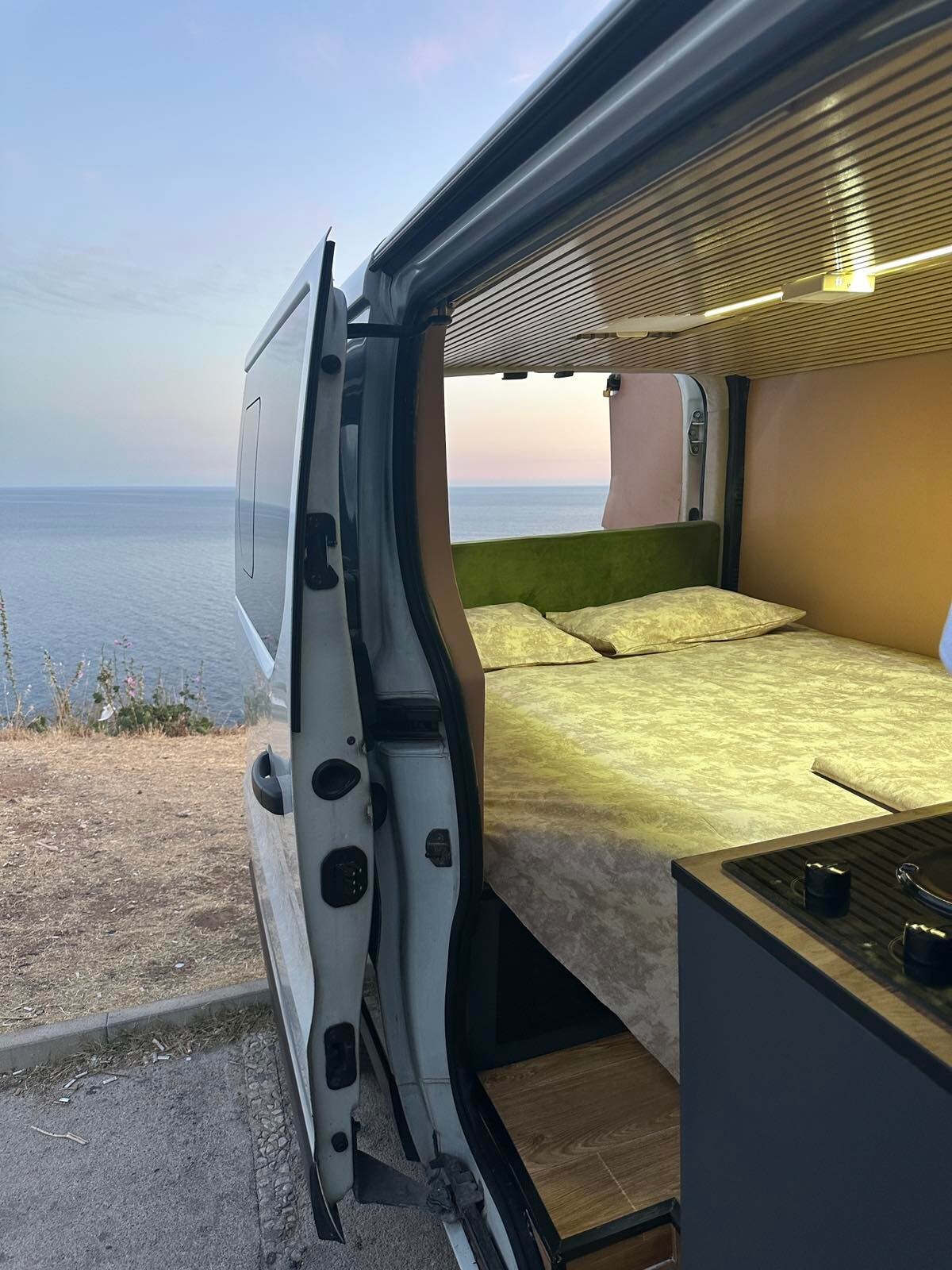 The interior of a camper van is visible, featuring a spacious bed with light-colored bedding. A small kitchenette area is equipped with a gas stove, and the open door frames an expansive view of the sea in the background, highlighting the tranquil setting.