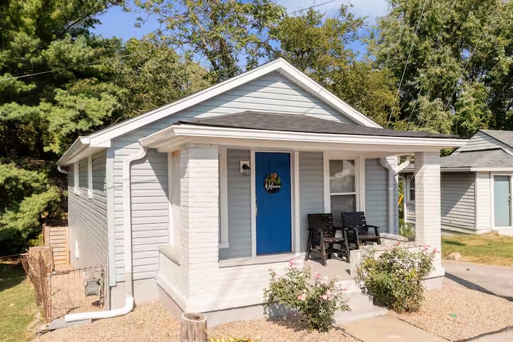 A charming house exterior features a vibrant blue front door, flanked by two black chairs on a covered porch. The light-colored exterior is surrounded by neatly manicured landscaping and gravel pathways, with greenery from nearby trees providing a natural backdrop.