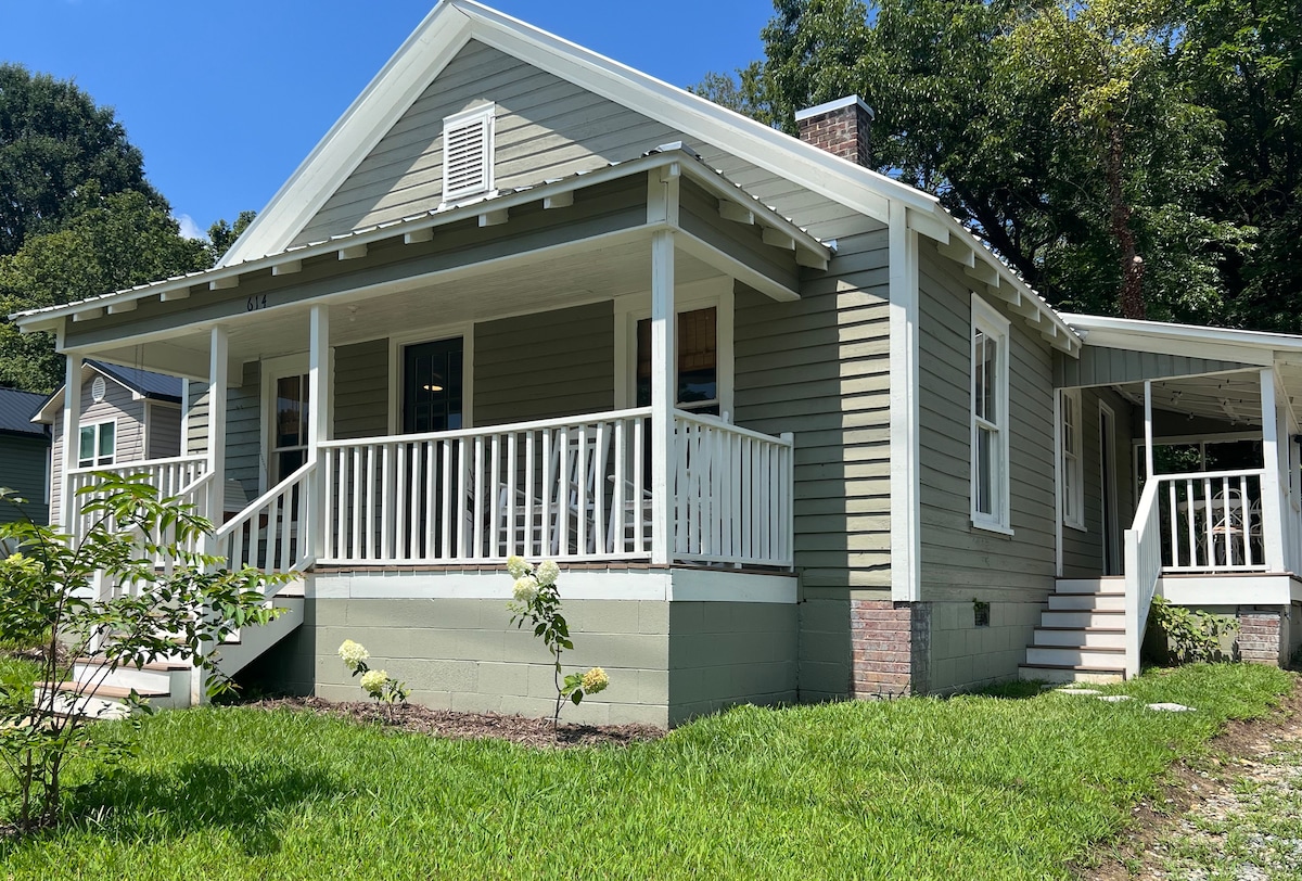 The exterior of a cozy cottage is displayed, featuring a light green façade and a classic gabled roof. A welcoming front porch with white railings and rocking chairs leads to a well-maintained yard, complete with lush green grass and flowering shrubs.