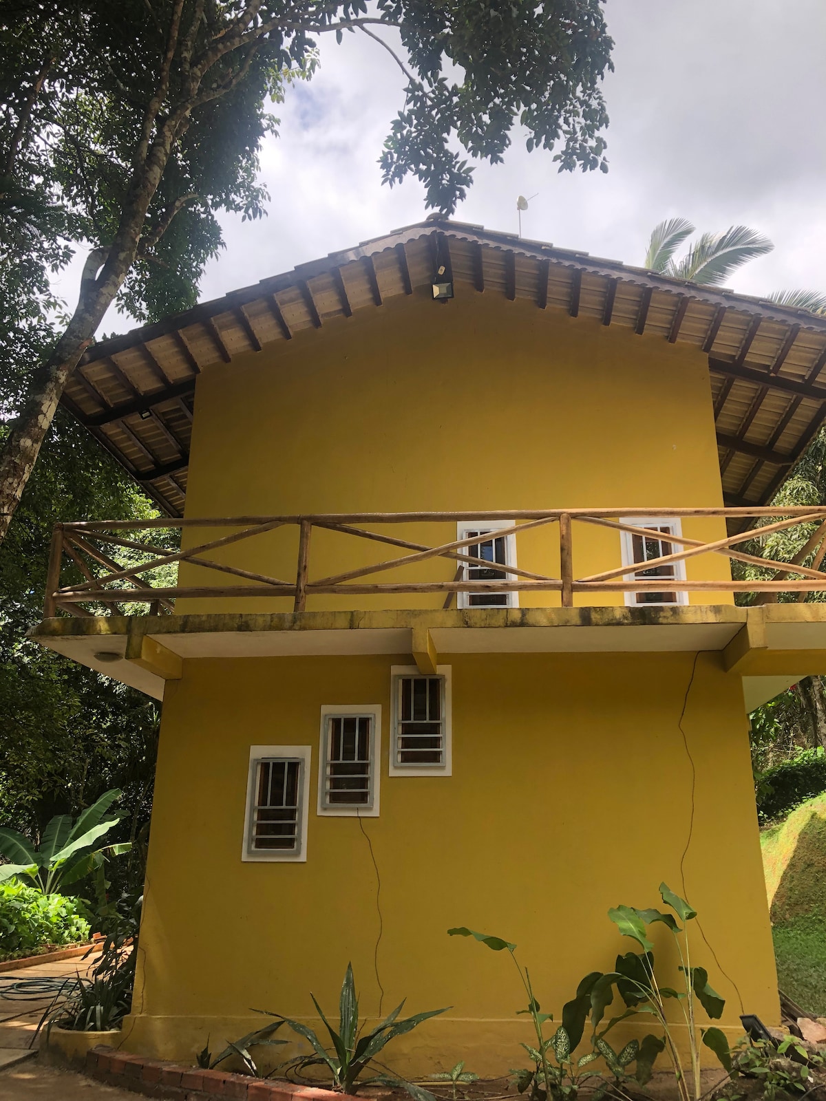 A two-story yellow house is surrounded by lush greenery. The balcony, supported by wooden railings, provides outdoor space on the upper level. Small windows are positioned along the side, allowing natural light to enter the interior. The sloped roof is adorned with wooden beams.