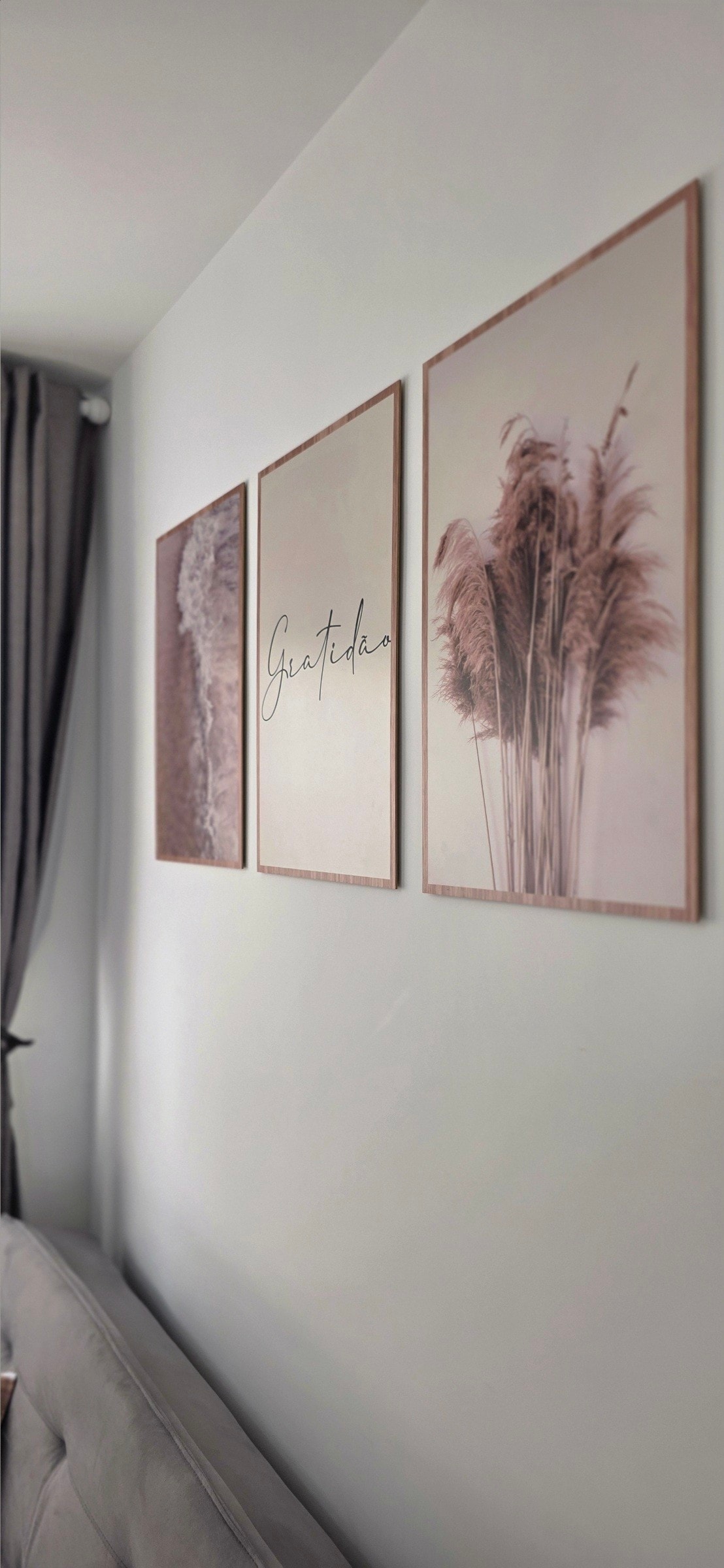 Three framed art pieces adorn a light-colored wall, featuring soft pampas grass imagery and elegant typography. A neutral-toned sofa is positioned in the foreground, complementing the modern decor. Sheer curtains allow natural light to filter softly into the space.