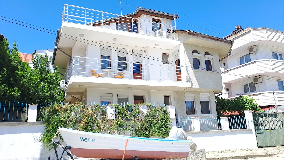 A two-story building is depicted, featuring a white exterior and a balcony with two chairs. Lush greenery surrounds the property, and a small boat, painted white, is positioned in the foreground. The sky above is clear and blue.