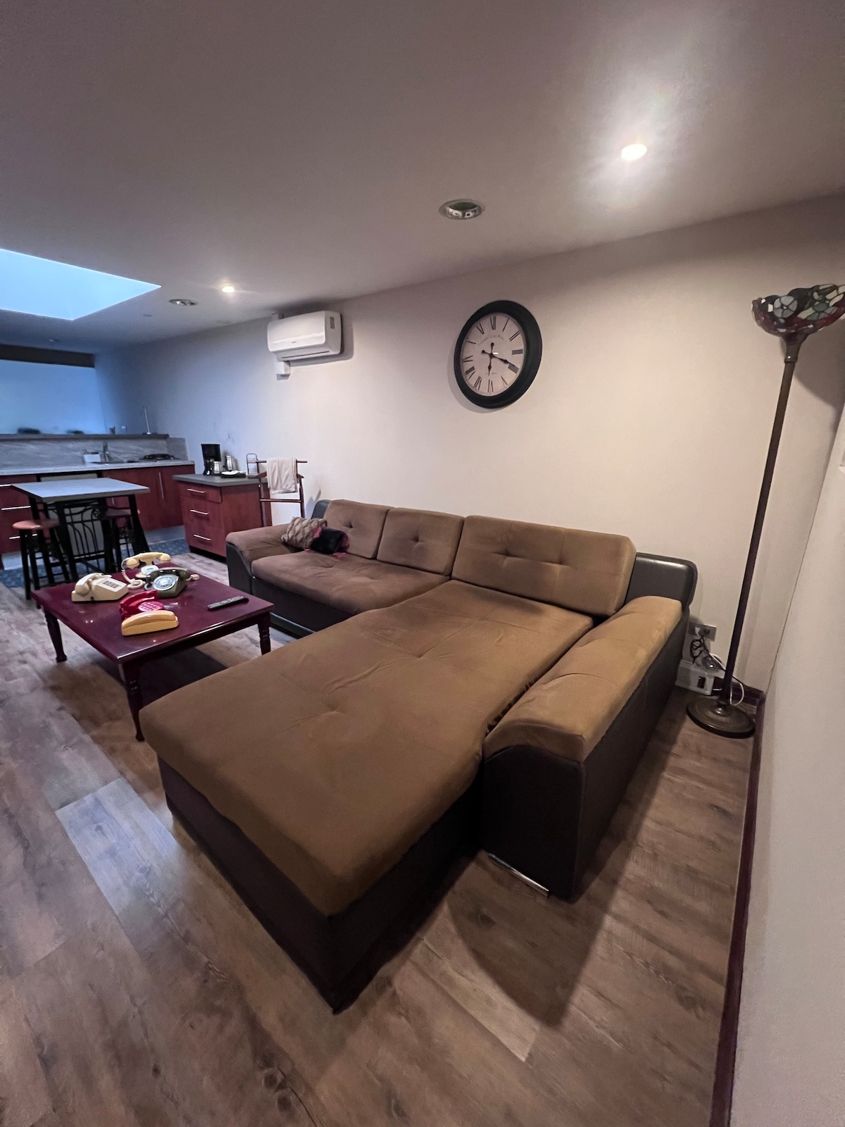 A cozy seating area is highlighted by a large sectional sofa in neutral tones. A coffee table holds various items, and a wall clock is mounted above. Warm lighting is provided by a nearby floor lamp, creating an inviting ambiance. The kitchen area can be seen in the background.
