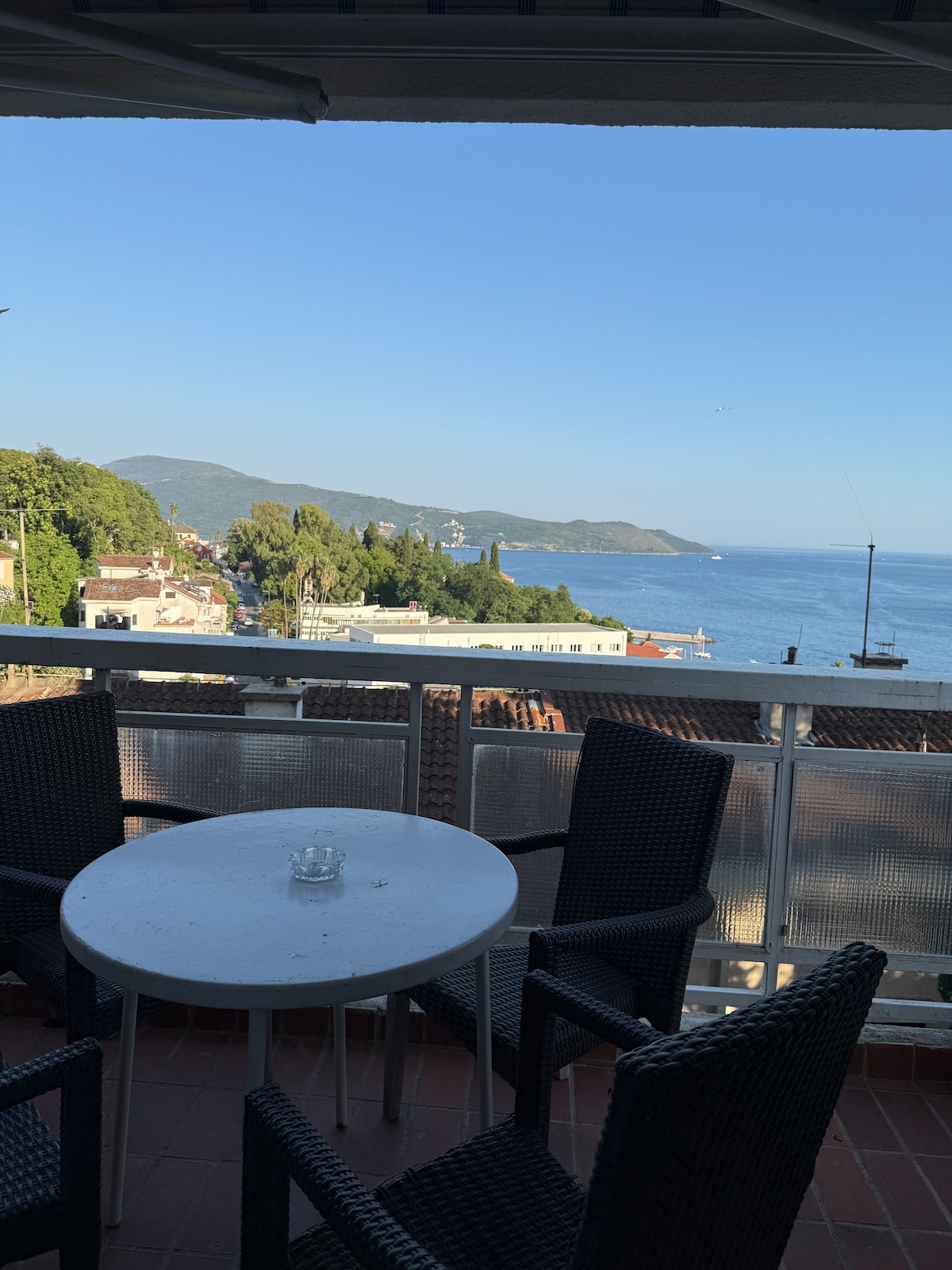 A private balcony overlooks the Adriatic Sea, showcasing a panoramic view of the shoreline and surrounding hills. A round table and four dark wicker chairs are arranged for outdoor seating, inviting guests to enjoy the serene atmosphere.