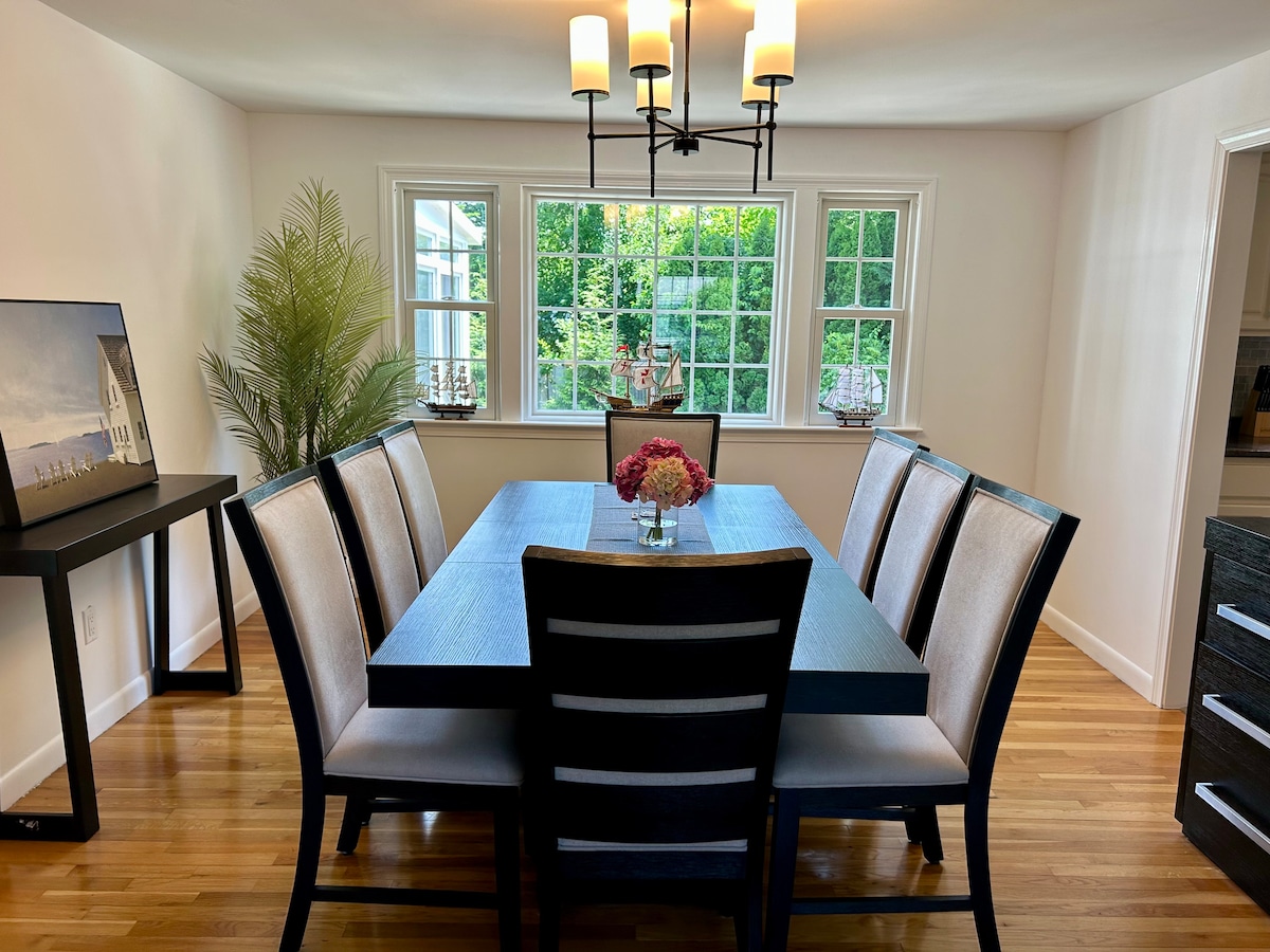 The dining room features a large table surrounded by eight upholstered chairs. Natural light enters through multiple windows, highlighting a floral centerpiece. A contemporary chandelier hangs above the table, and a side console complements the space.